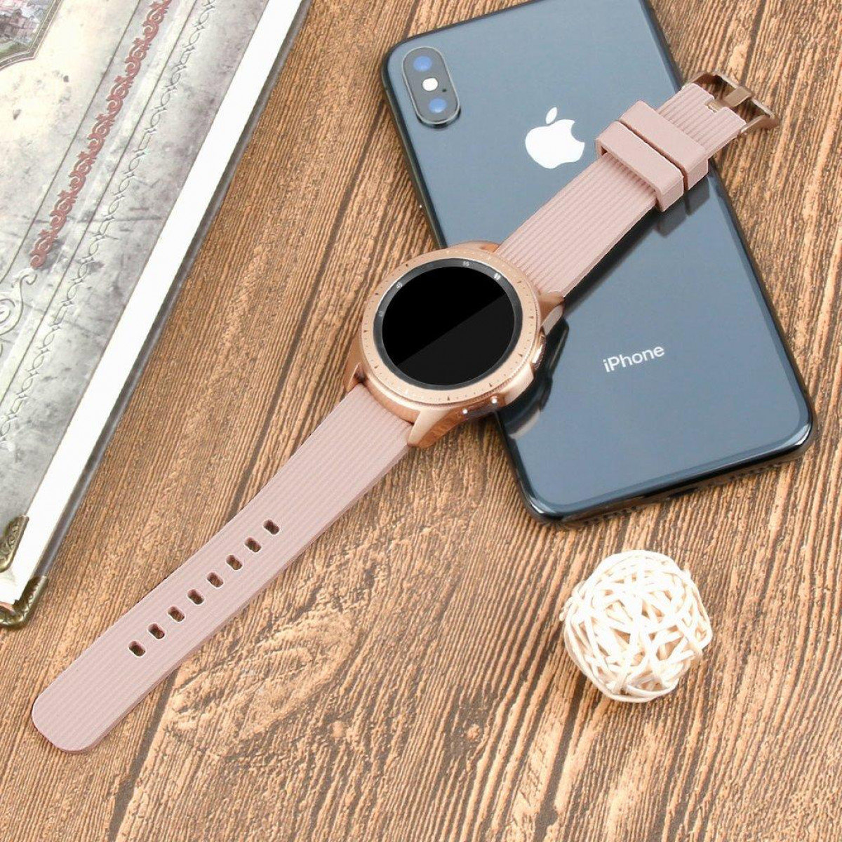 Roségoldene Smartwatch mit rosa Armband, neben einem schwarzen iPhone auf Holzoberfläche, mit weißem Ball.