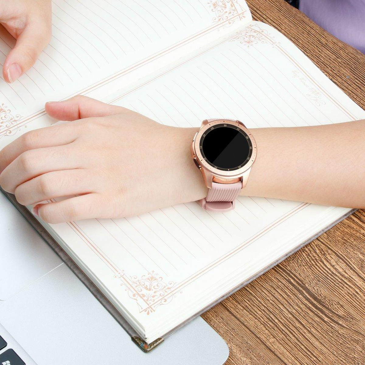 Die Hand einer Person mit einer roségoldenen Smartwatch auf einem Notizbuch und einem Holztisch.