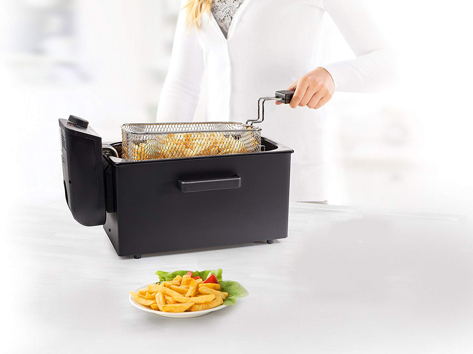 Kobieta wyjmuje kosz frytek z czarnej frytkownicy. Talerz frytek z przodu.