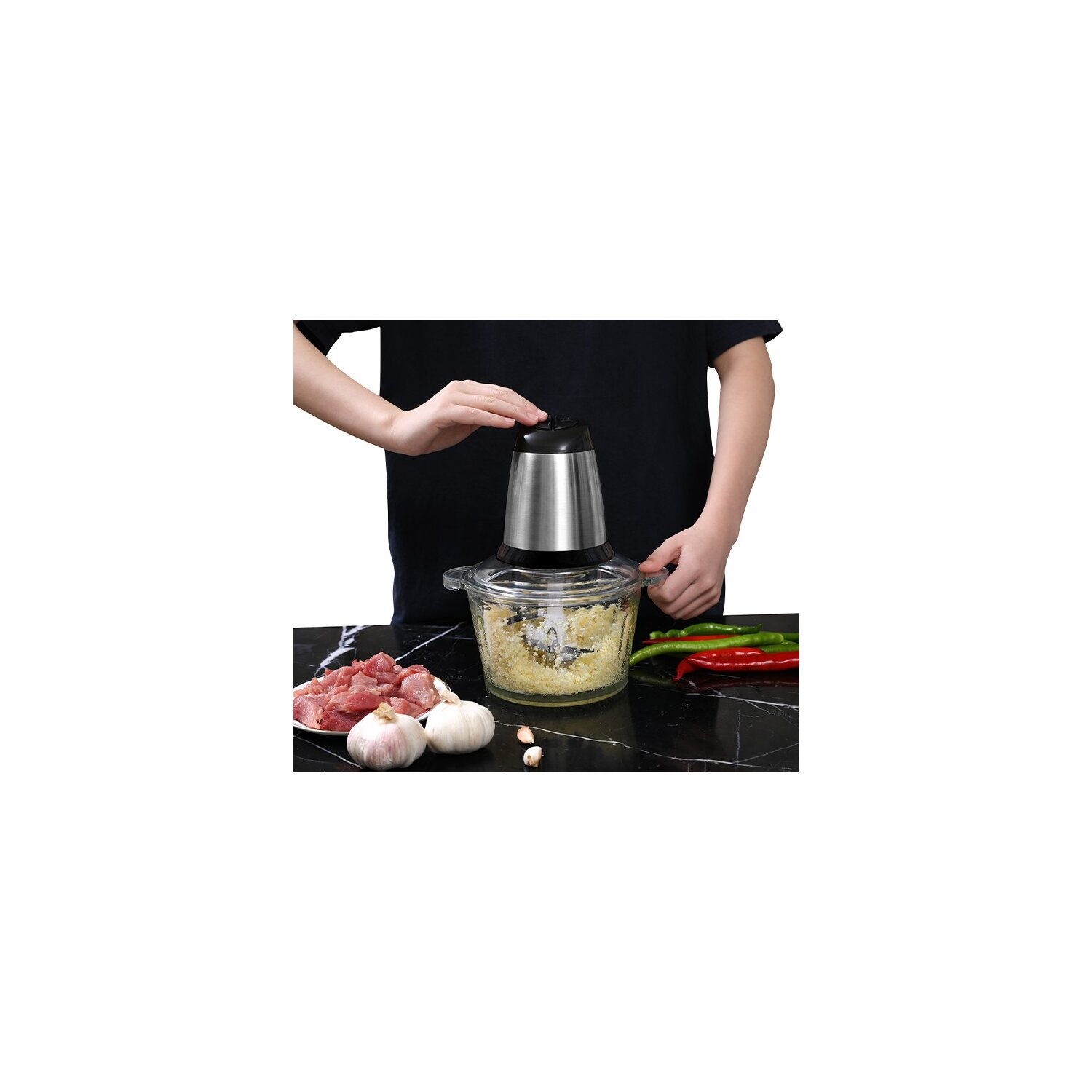 Eine Person benutzt eine silberne Küchenmaschine. Knoblauch, Fleisch und Paprika sind auf dem schwarzen Tisch zu sehen.
