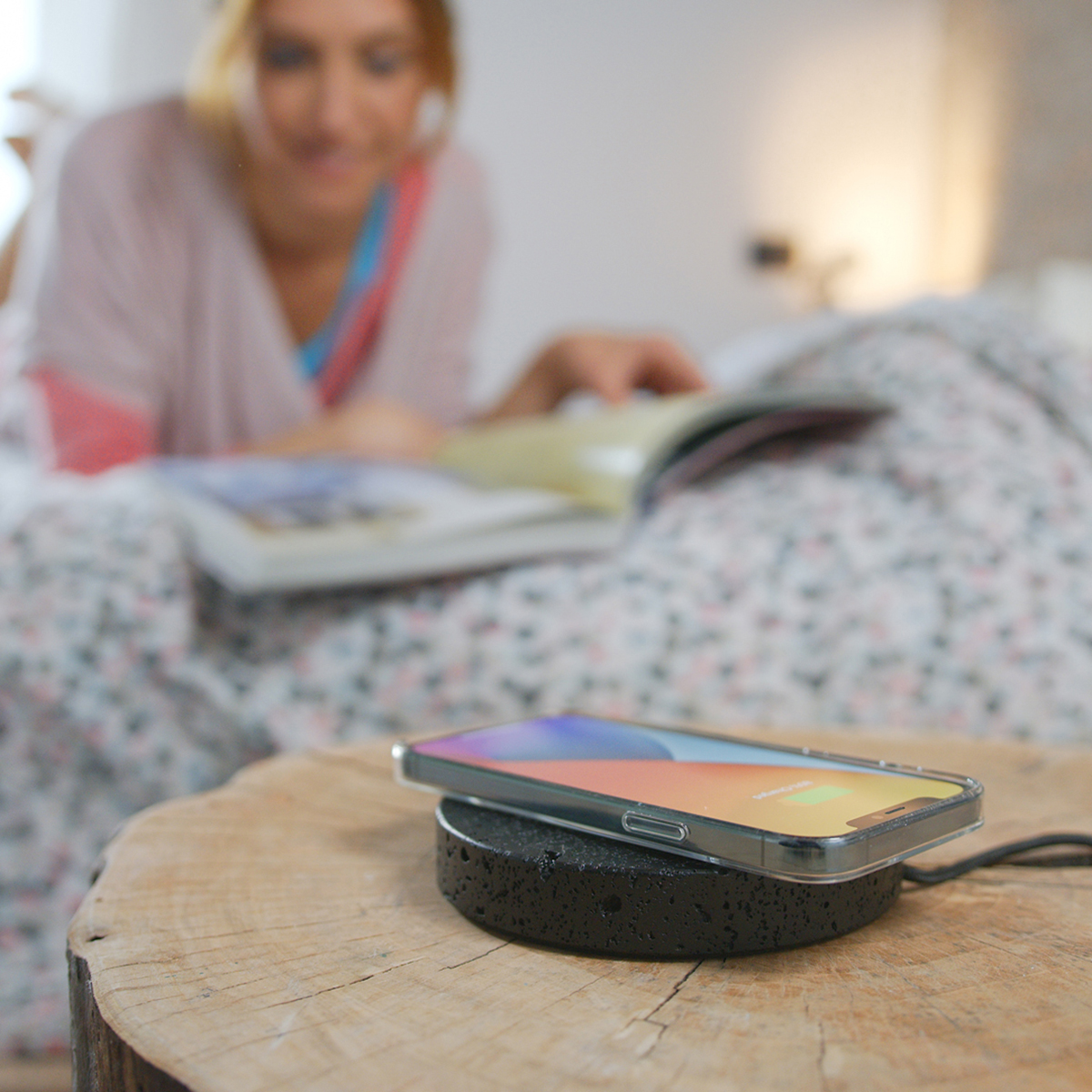 Ein Telefon, das auf einem schwarzen kabellosen Ladegerät auf einem Holztisch geladen wird. Im Hintergrund liest eine Frau.