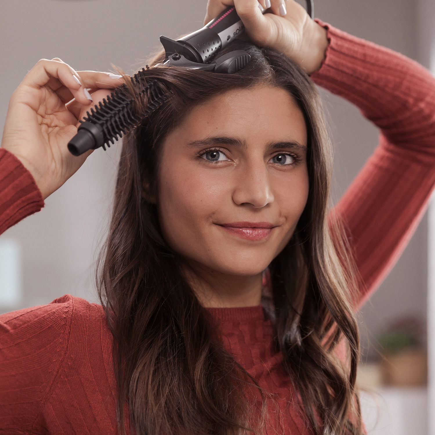 Una mujer usa un cepillo para rizar el cabello, sonriendo. El fondo está borroso.