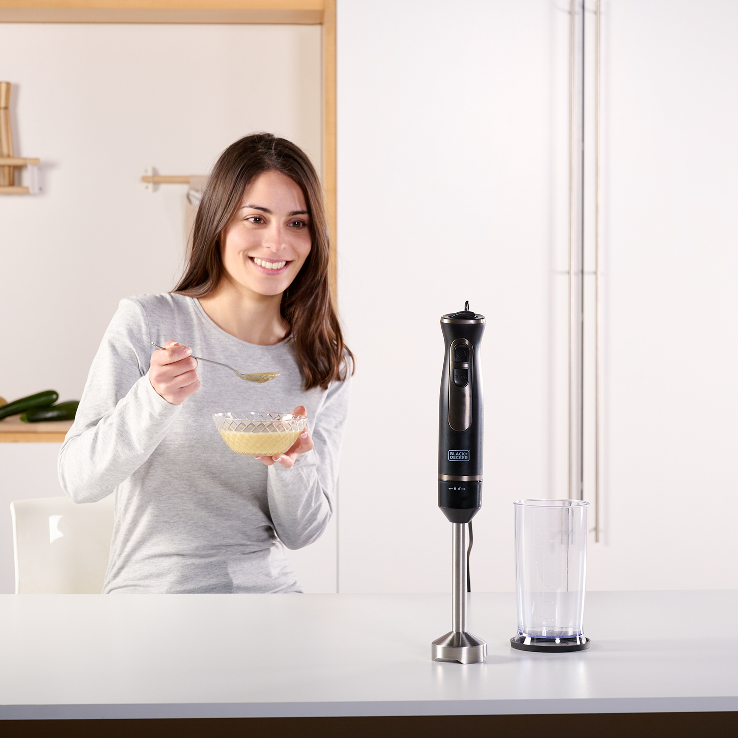 Kobieta uśmiecha się, trzymając miskę z zupą i łyżkę. Na stole stoi blender i szklanka.