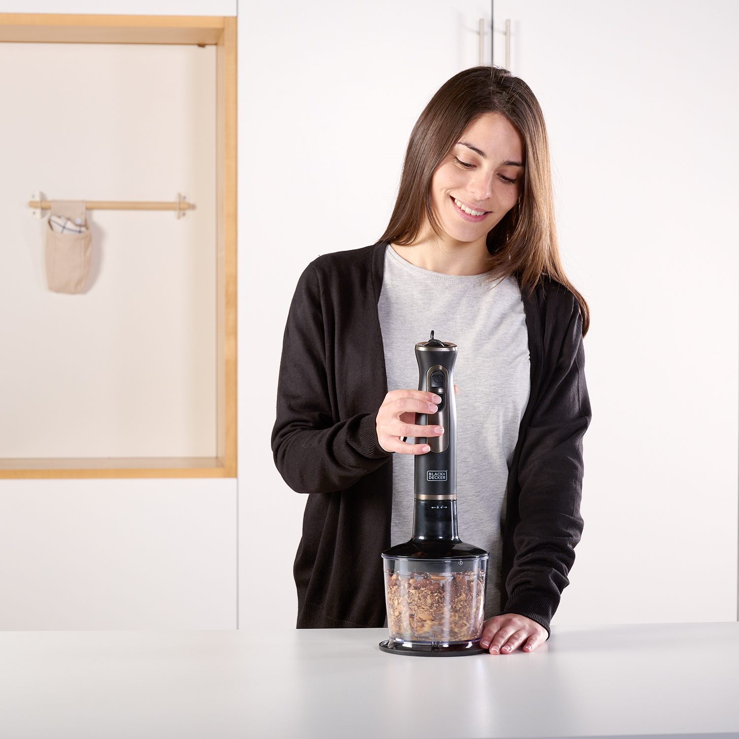 Kobieta trzyma czarny blender nad pojemnikiem z jedzeniem na białej powierzchni. Tło to biała kuchnia.