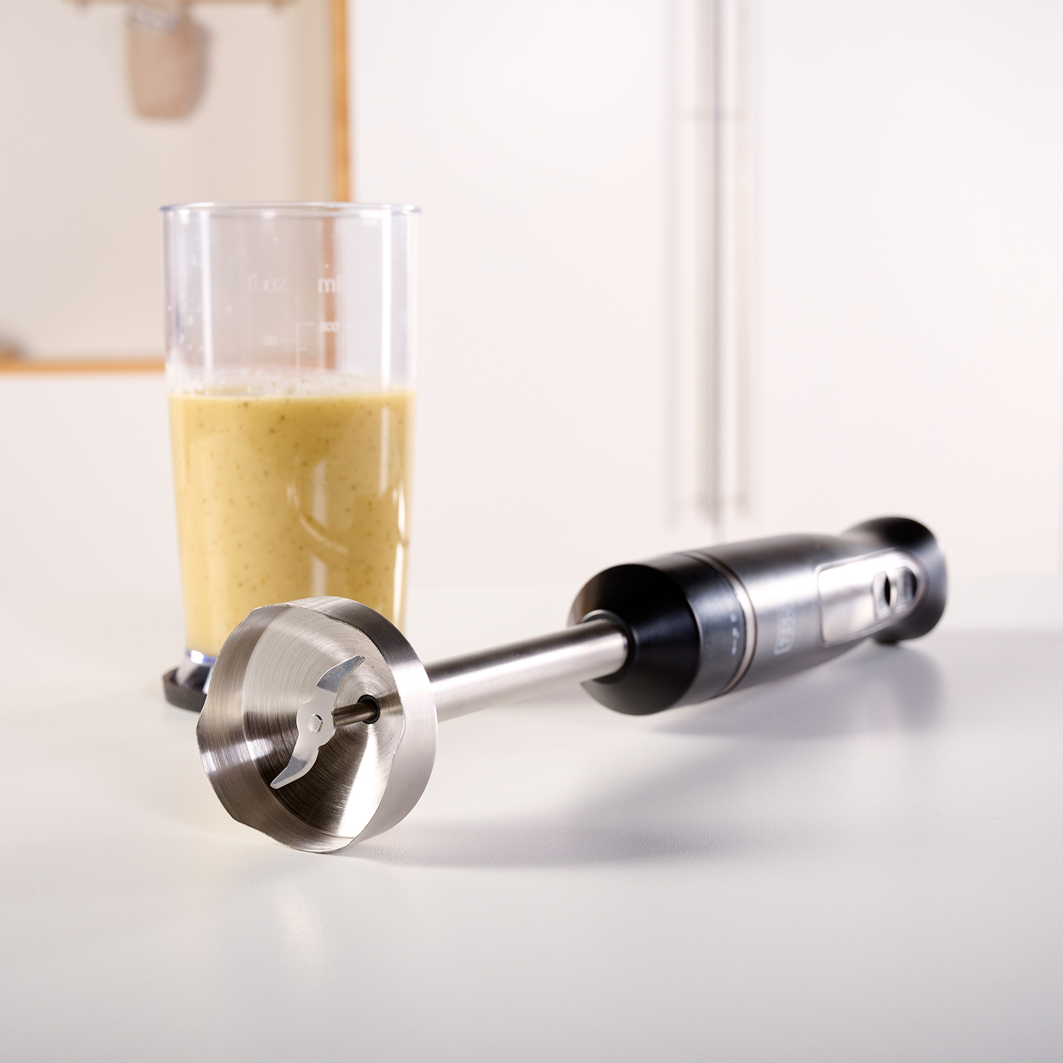 Blender ze smoothie w przezroczystym pojemniku. Blender jest czarny i srebrny. Stoi na białej powierzchni.