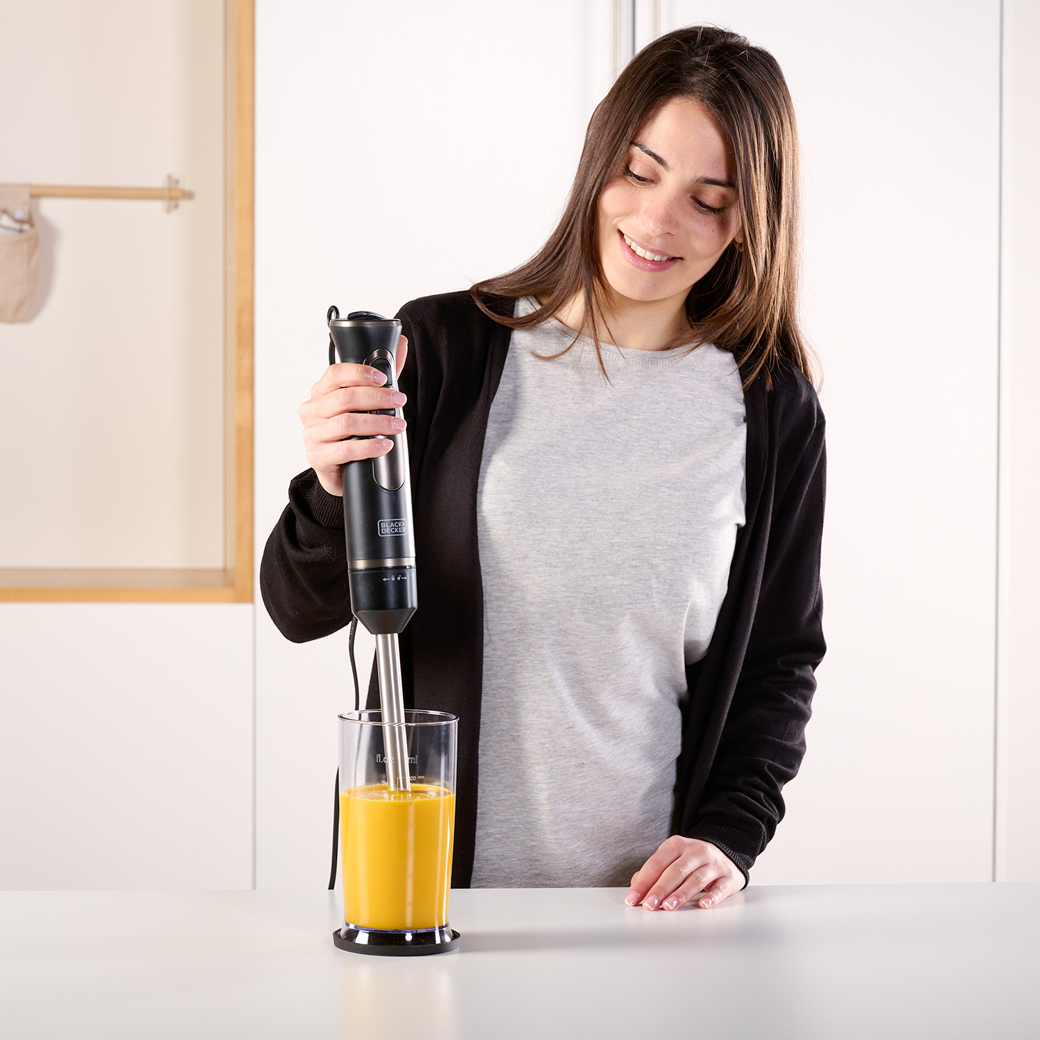 Kobieta używa czarnego blendera ręcznego w szklance z pomarańczowym płynem.