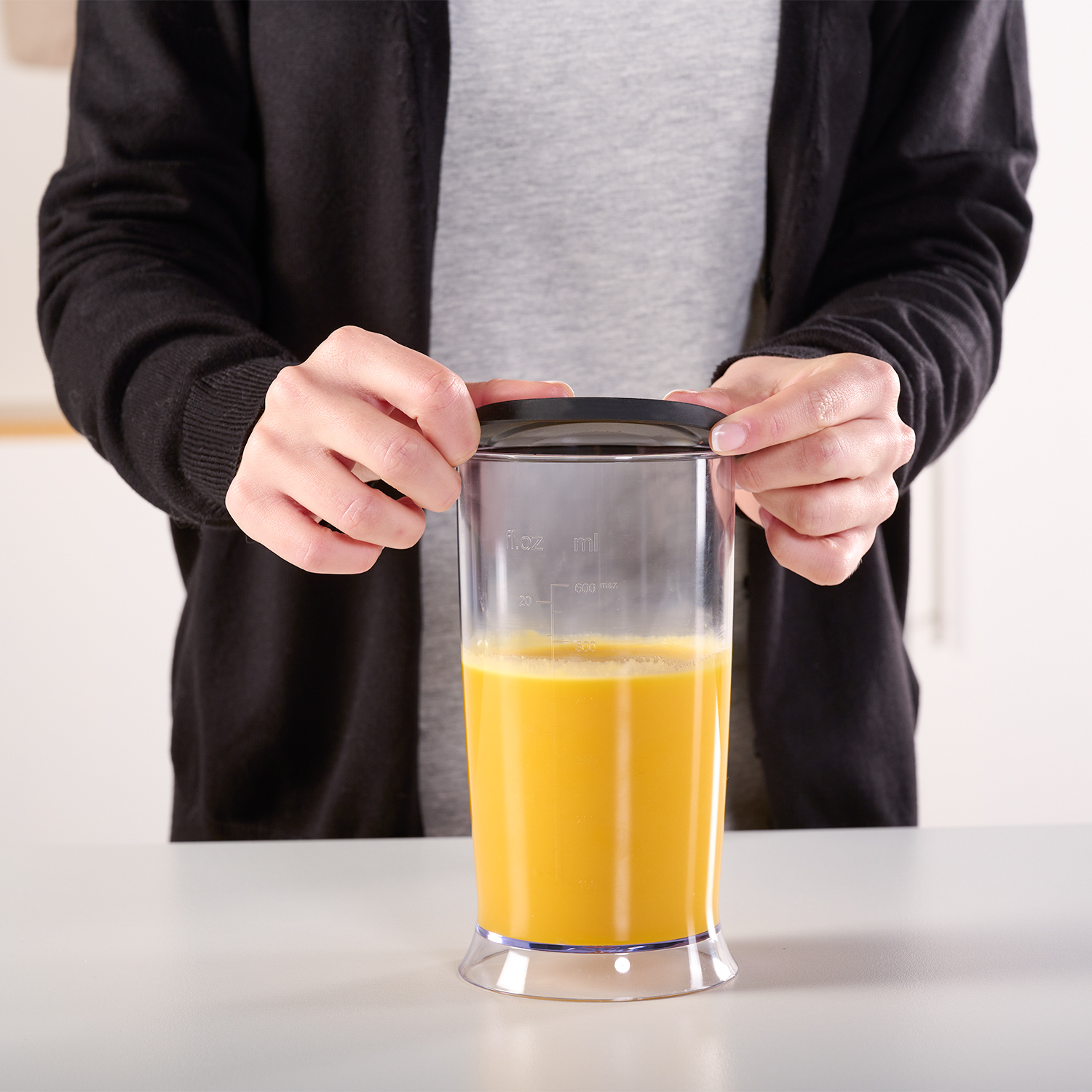 Ręce trzymające przezroczysty pojemnik blendera z sokiem pomarańczowym na białej powierzchni.