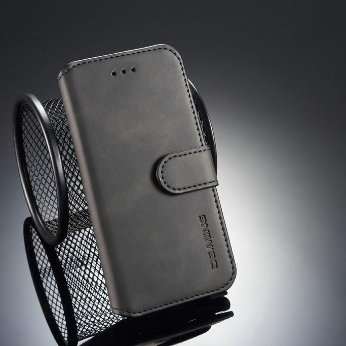 Czarny futerał na telefon z zapięciem, trzymający telefon, umieszczony obok czarnego pojemnika z siatki na odblaskowej powierzchni.