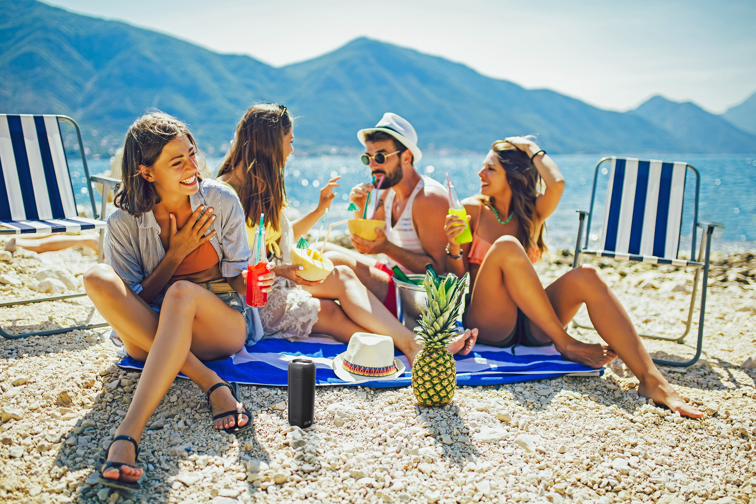 Scena sulla spiaggia. Quattro persone con bevande e un altoparlante. Montagne e mare sullo sfondo.