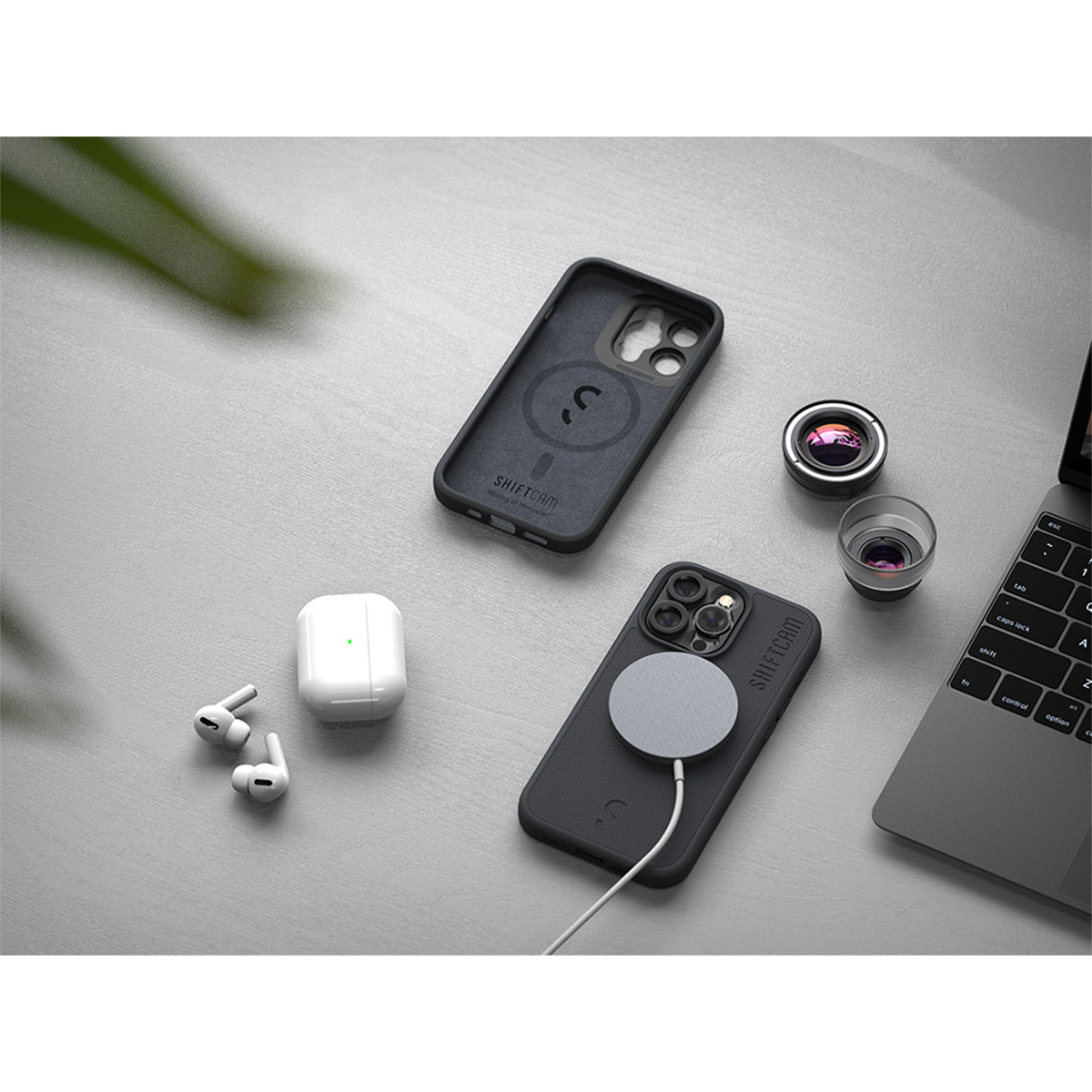 Telefon mit Hülle, AirPods, Linse und Laptop auf einem Schreibtisch. Artikel sind ordentlich angeordnet.
