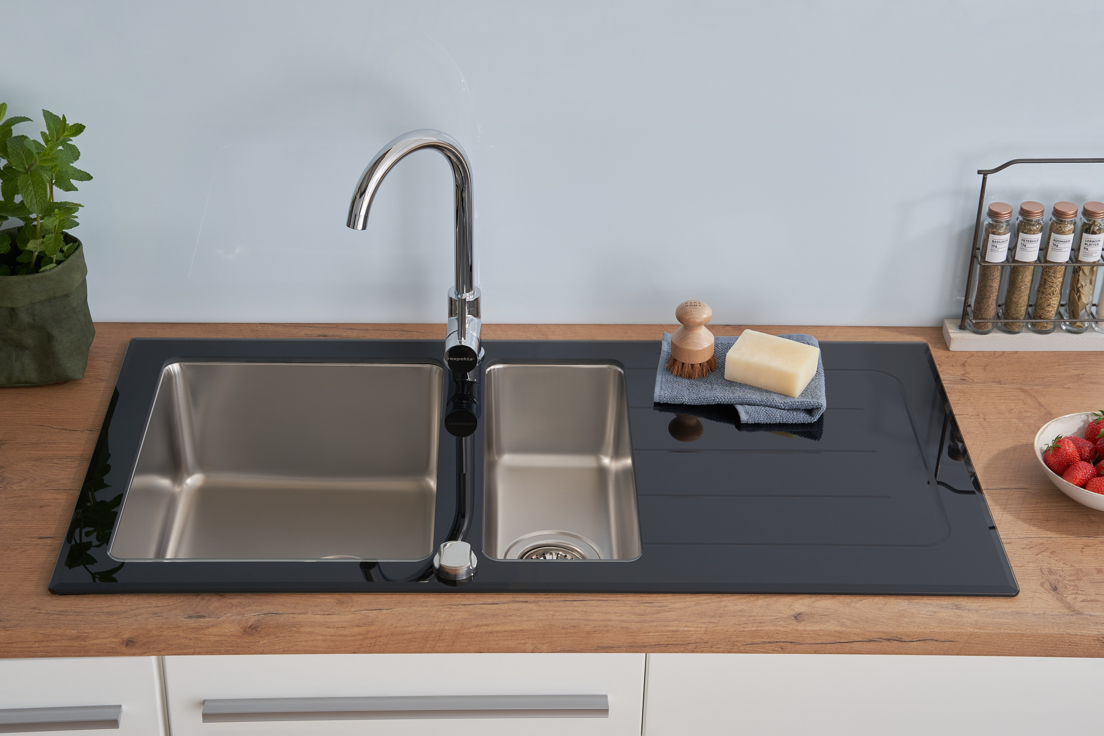 Edelstahl-Küchenspüle mit schwarzer Arbeitsplatte, Wasserhahn und Spülbürste. Erdbeeren und Kräuter sind in der Nähe.