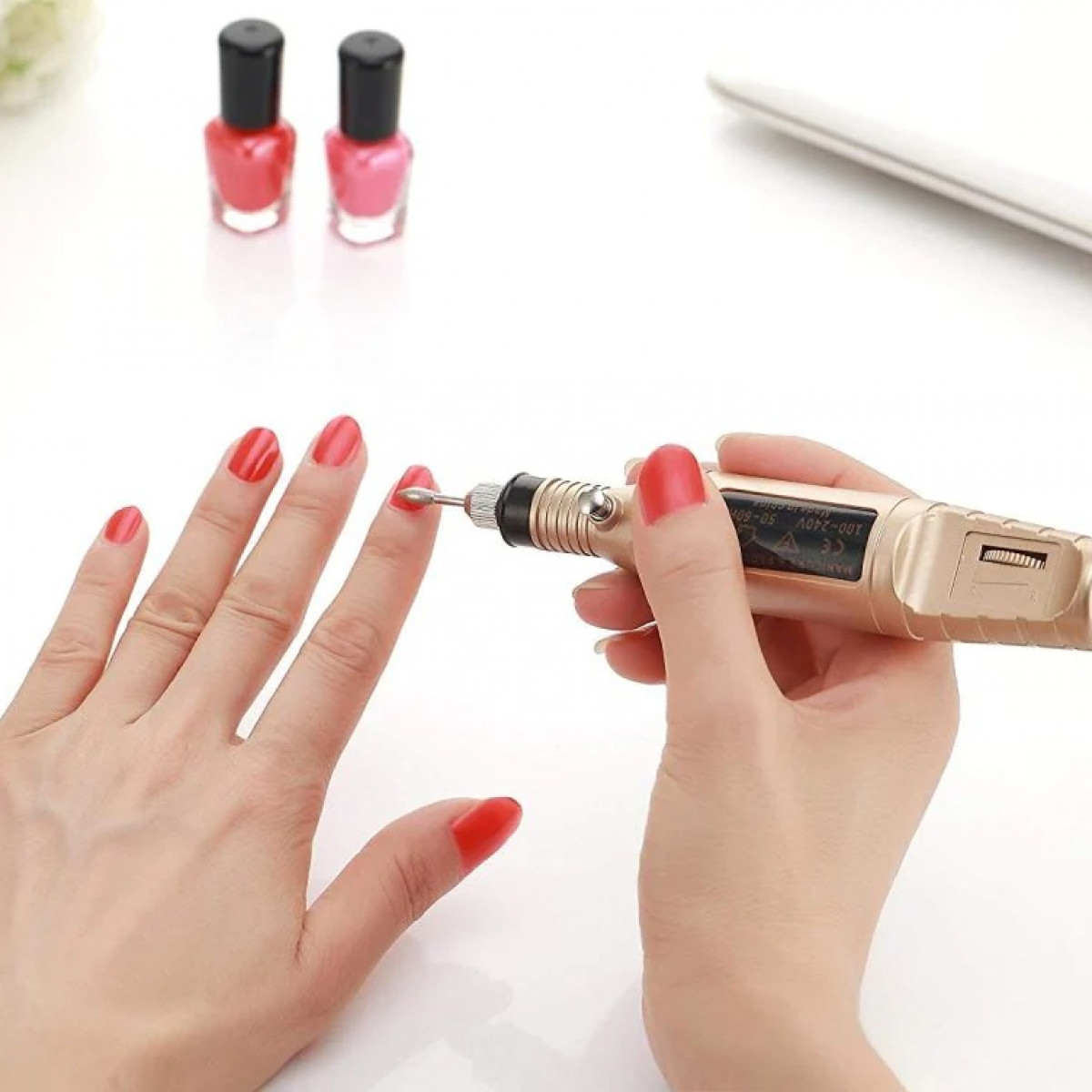 Kobieta używa frezarki do paznokci na pomalowanym na czerwono paznokciu. Dwa słoiczki lakieru do paznokci w tle.