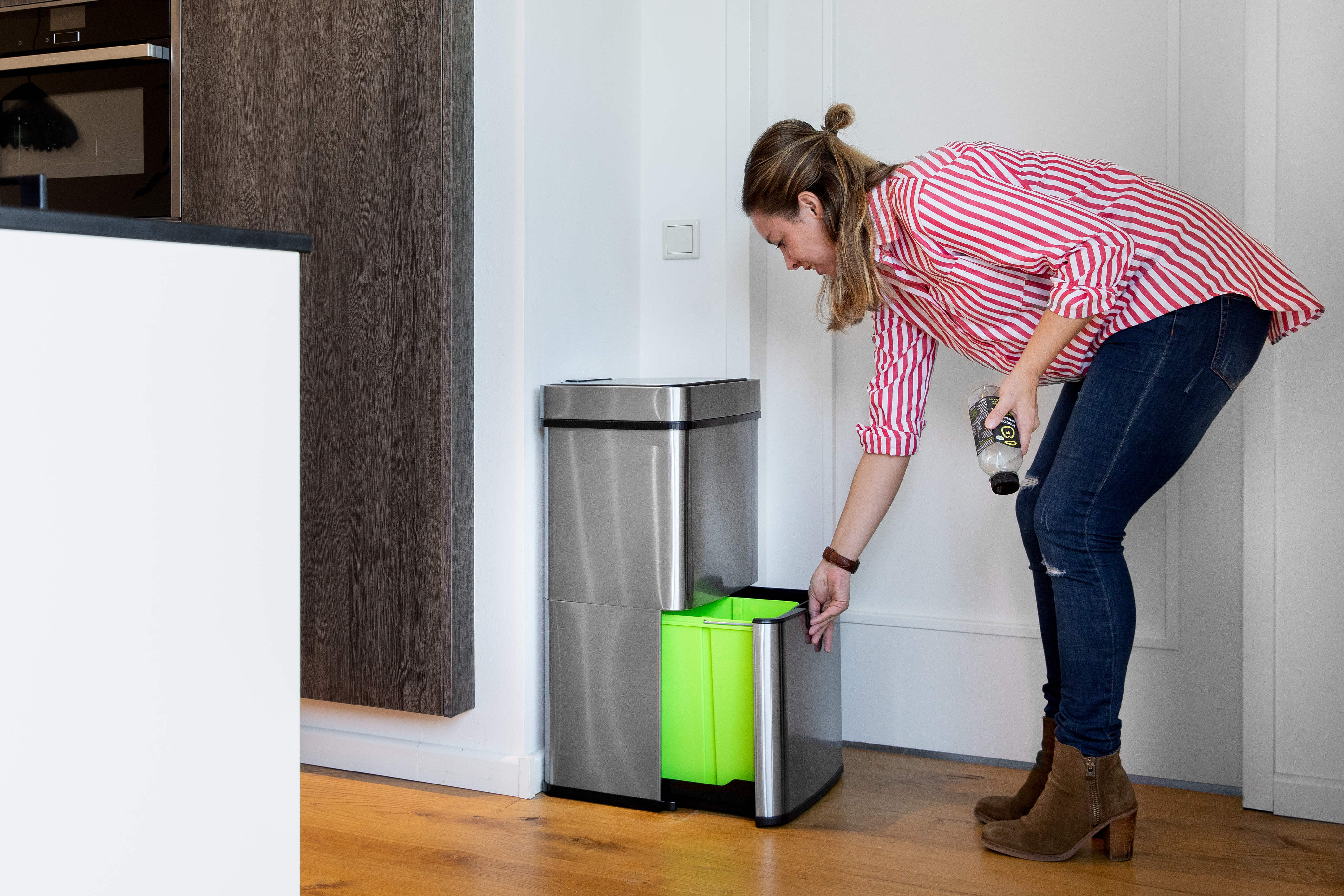 Eine Frau wirft eine Plastikflasche in einen grünen Mülleimer. Küchenhintergrund.