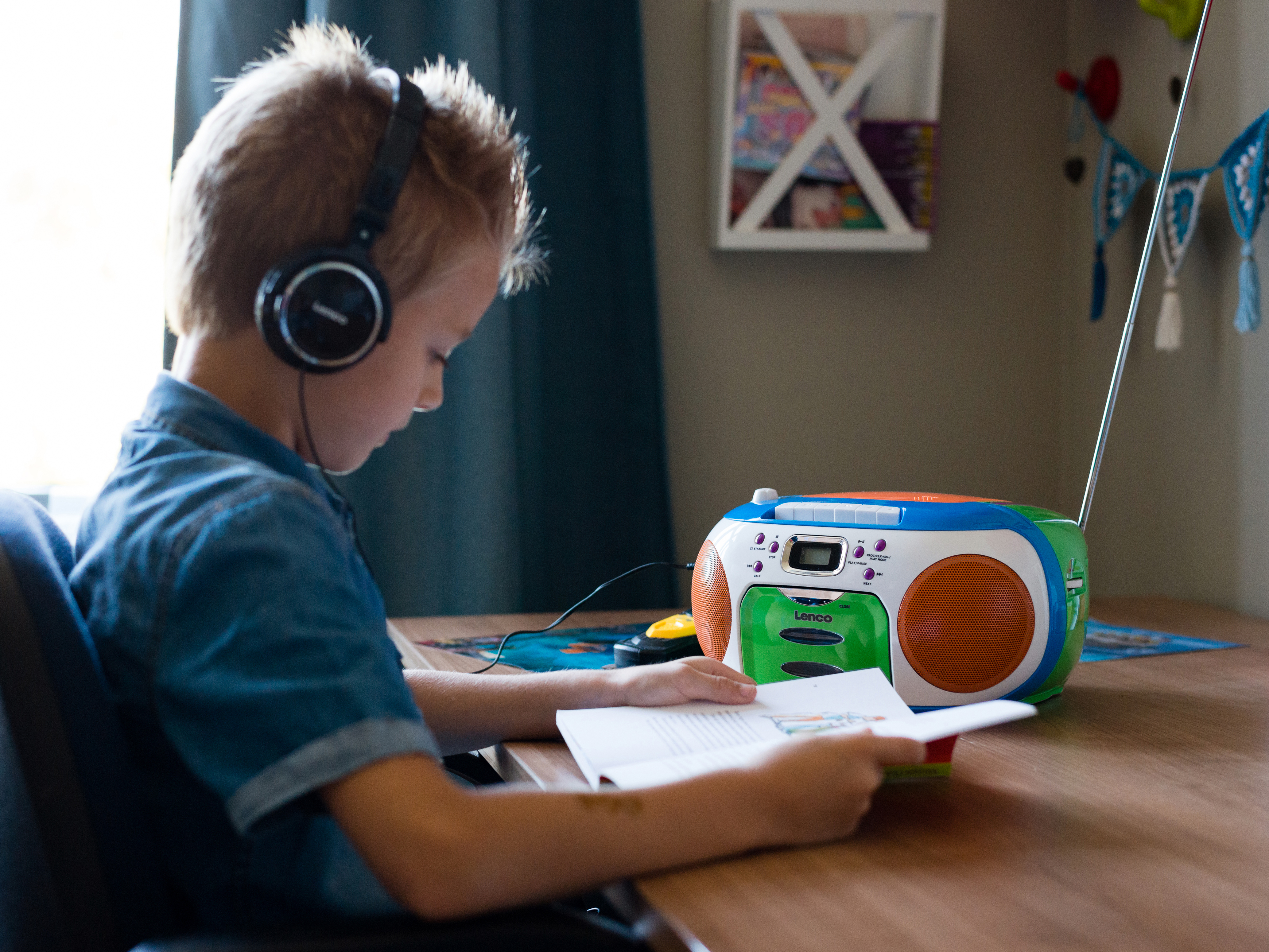 Un niño con auriculares lee un libro en una mesa. Una radio y decoraciones están en el fondo.