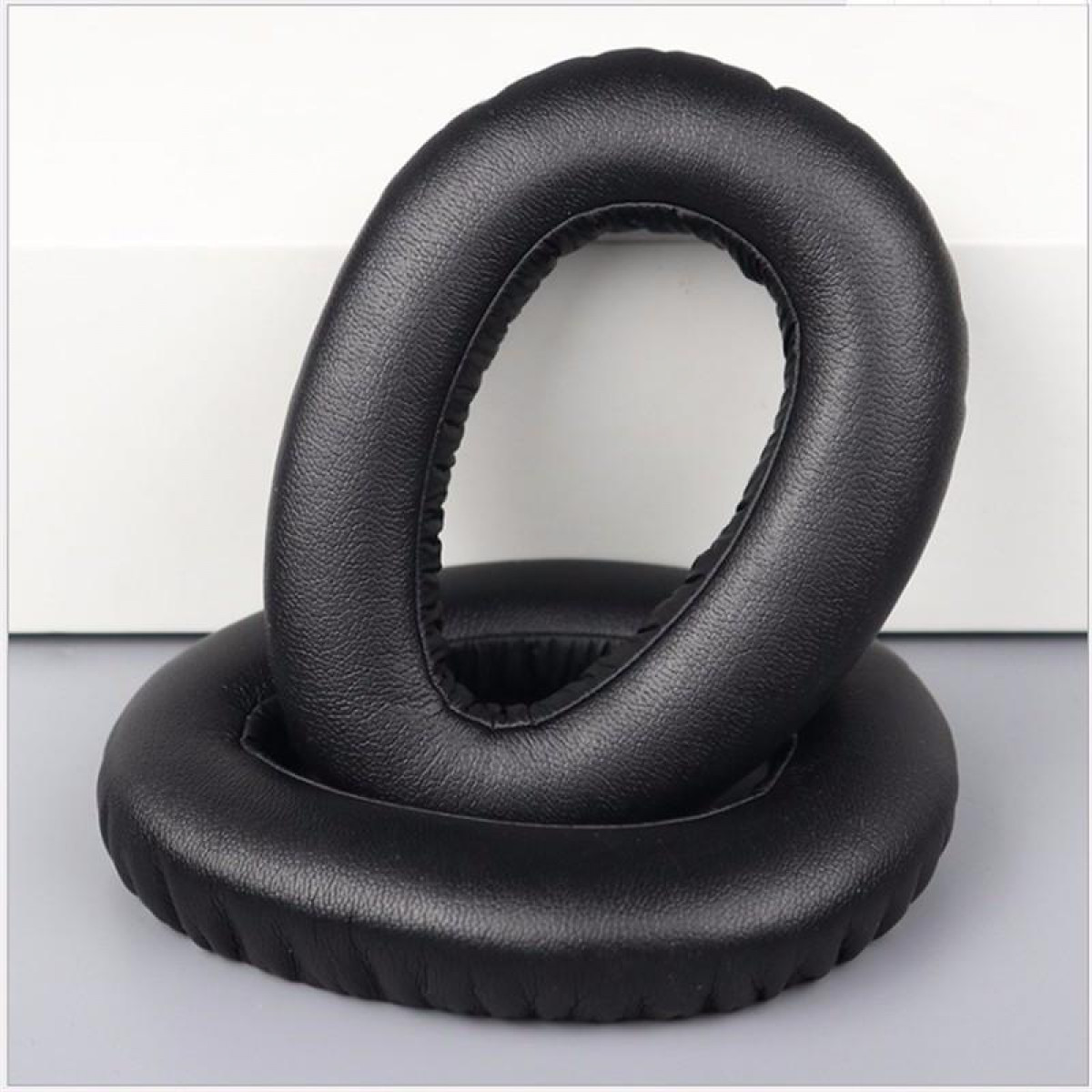 Dwie czarne, owalne poduszki na uszy. Leżą na jasnoszarej powierzchni.