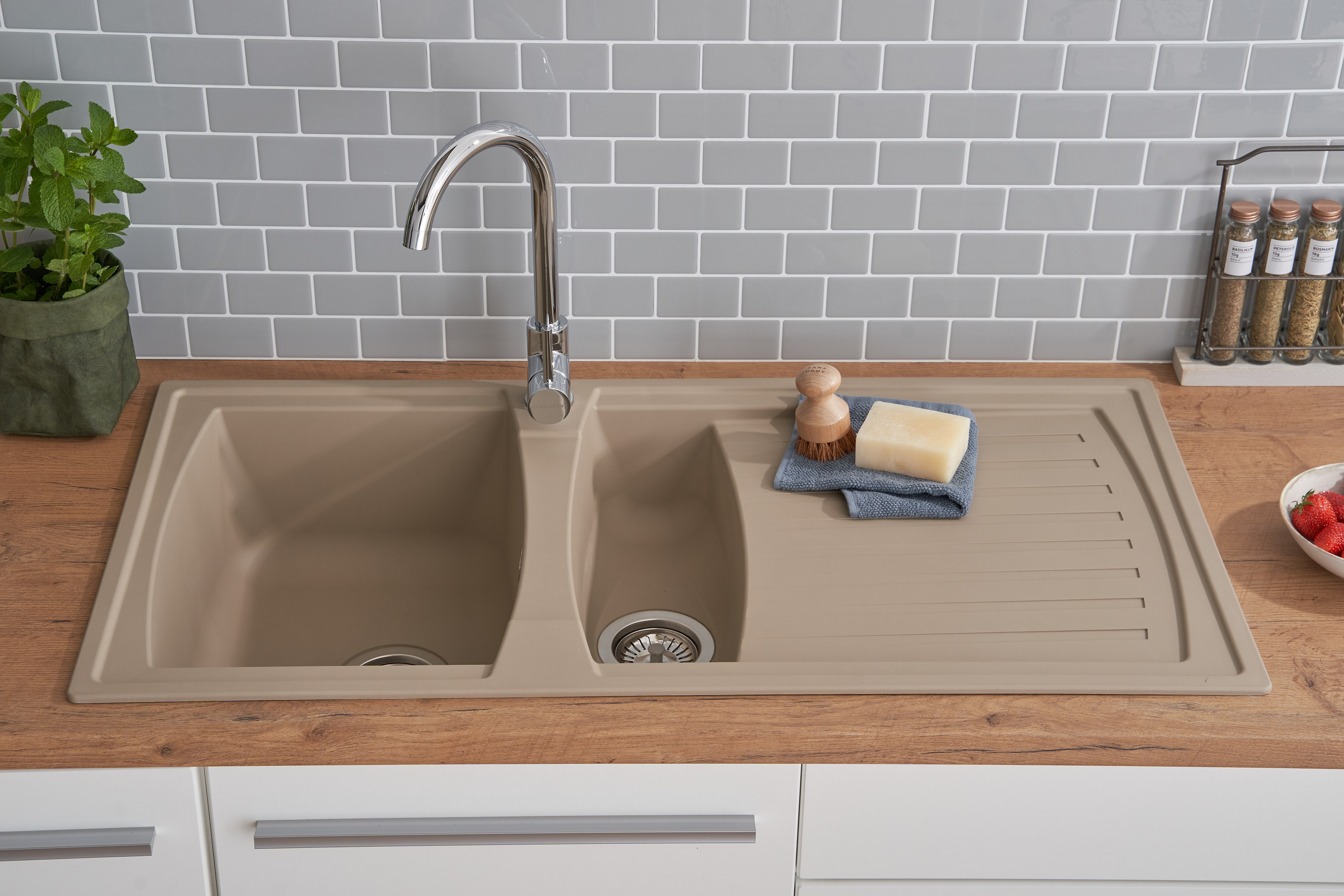Ein Küchenbecken mit Wasserhahn, Seife, Bürste und grauer Fliesenrückwand auf einer Holz-Arbeitsplatte.