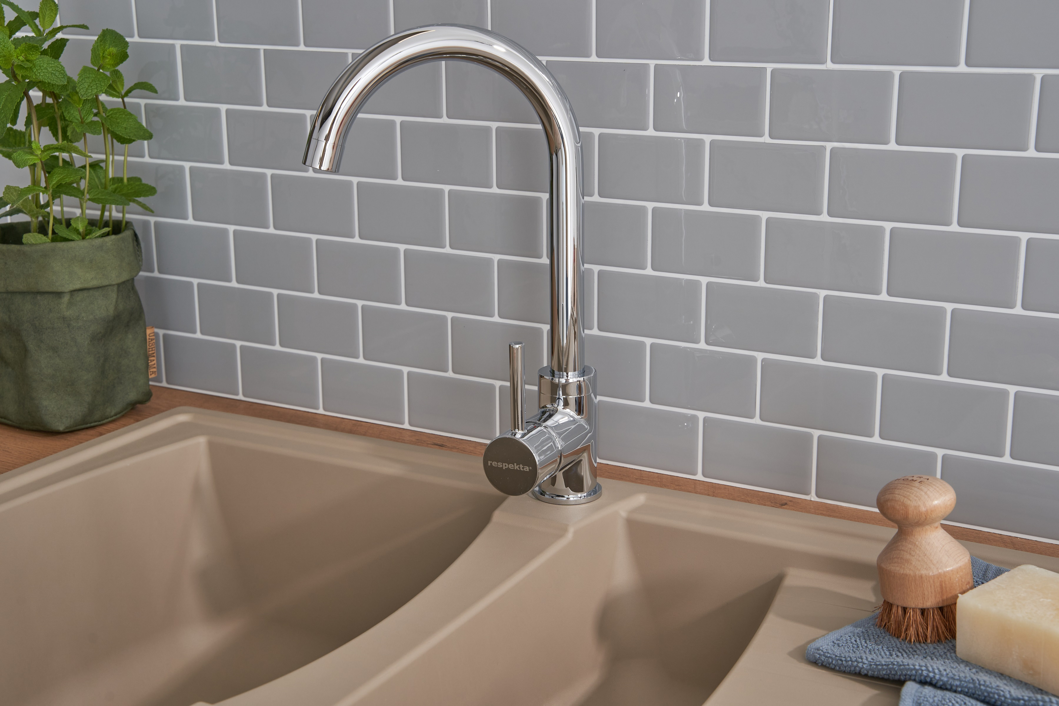 Chrom-Wasserhahn über einem beigen Waschbecken vor einer grau gefliesten Wand. Eine Pflanze und eine Bürste sind vorhanden.