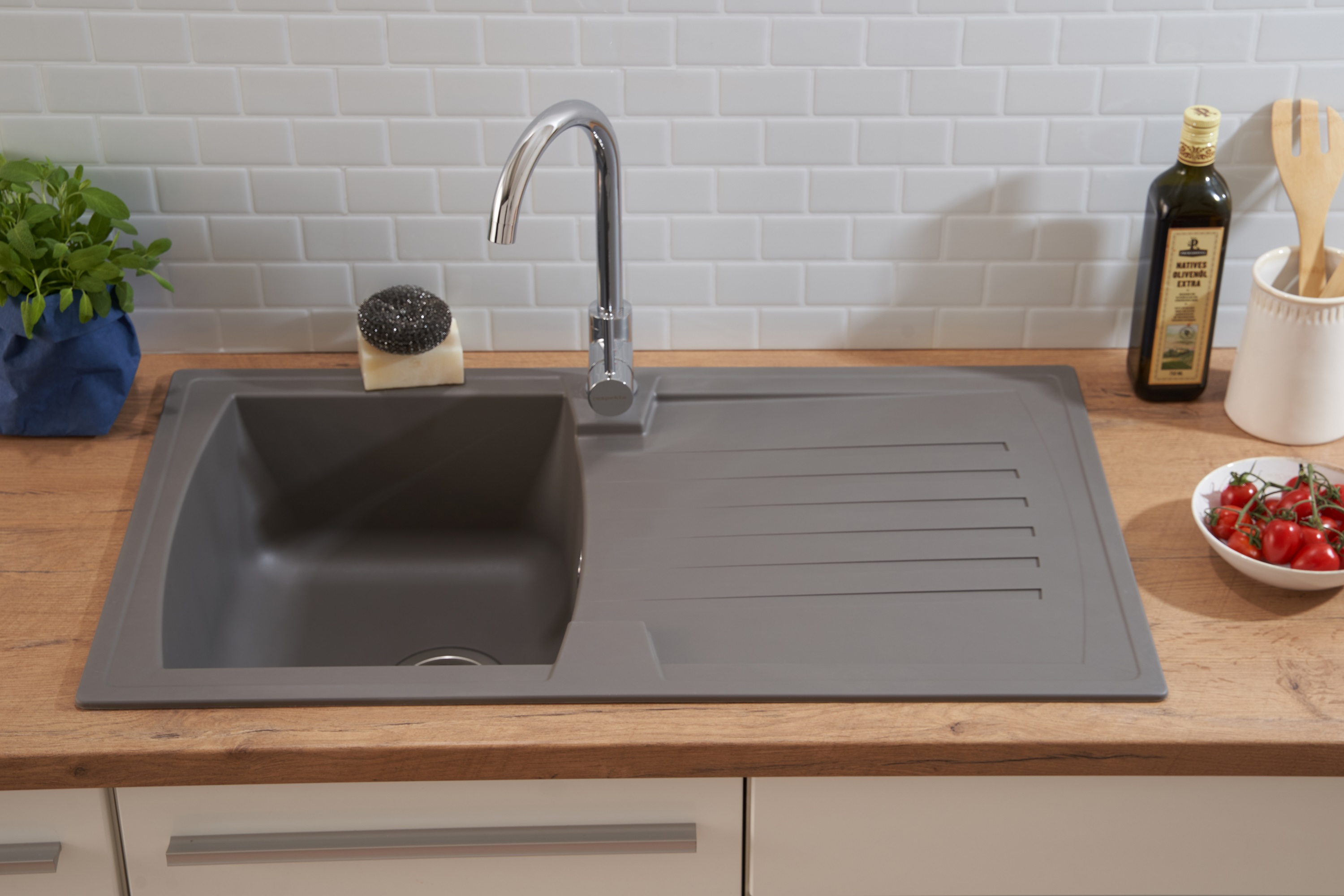Graue Küchenspüle, Wasserhahn, Holztisch, Olivenöl, Tomaten und ein Schwamm.