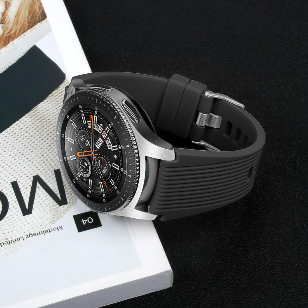 Silberne und schwarze Smartwatch mit schwarzem Armband, auf weißem Magazin. Dunkler Hintergrund.