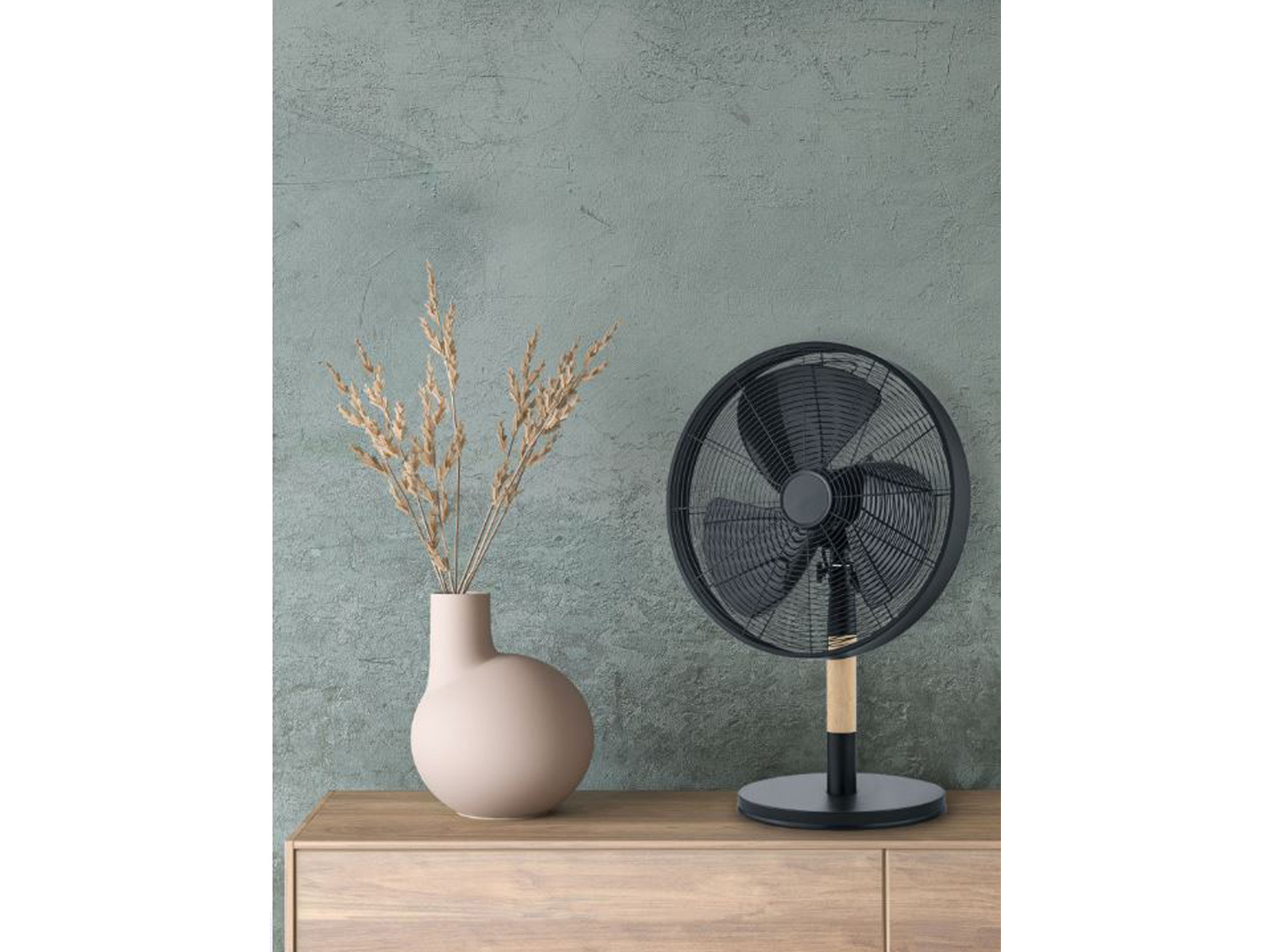 Ein schwarzer Ventilator auf einem Holzschrank, beige Vase mit Pflanzen, grüner Wandhintergrund.