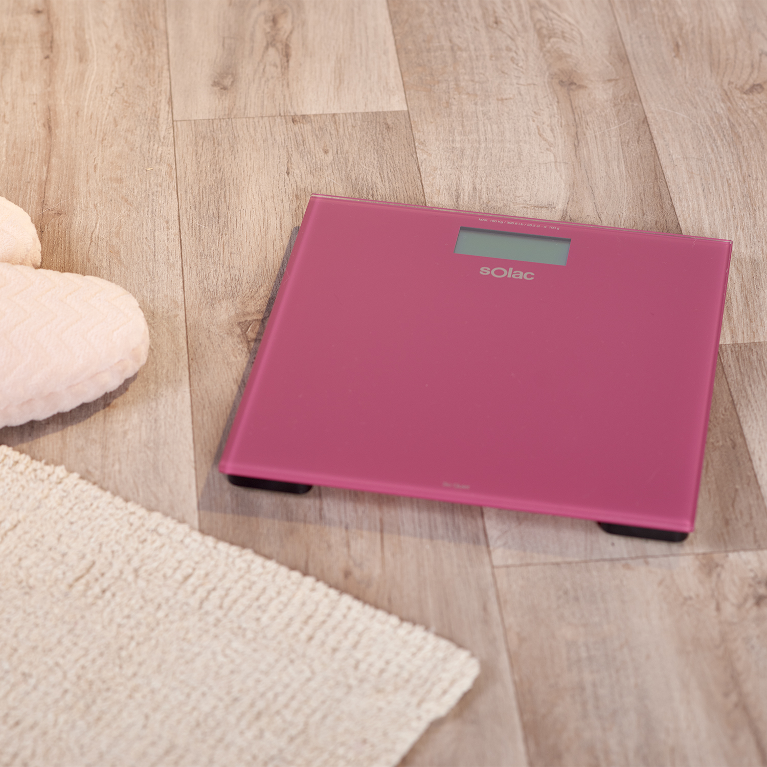 Eine rosa Digitalwaage steht auf einem Holzboden. Ein beiger Teppich und ein rosa Kissen sind in der Nähe.