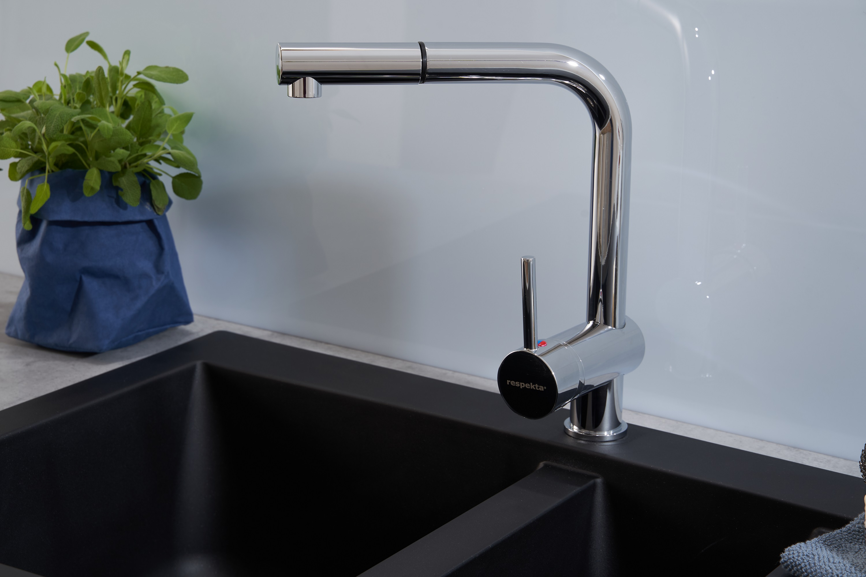 Ein Chrom-Wasserhahn mit einem schwarzen Waschbecken. Daneben befindet sich eine Pflanze in einer blauen Tüte. Weißer Hintergrund.