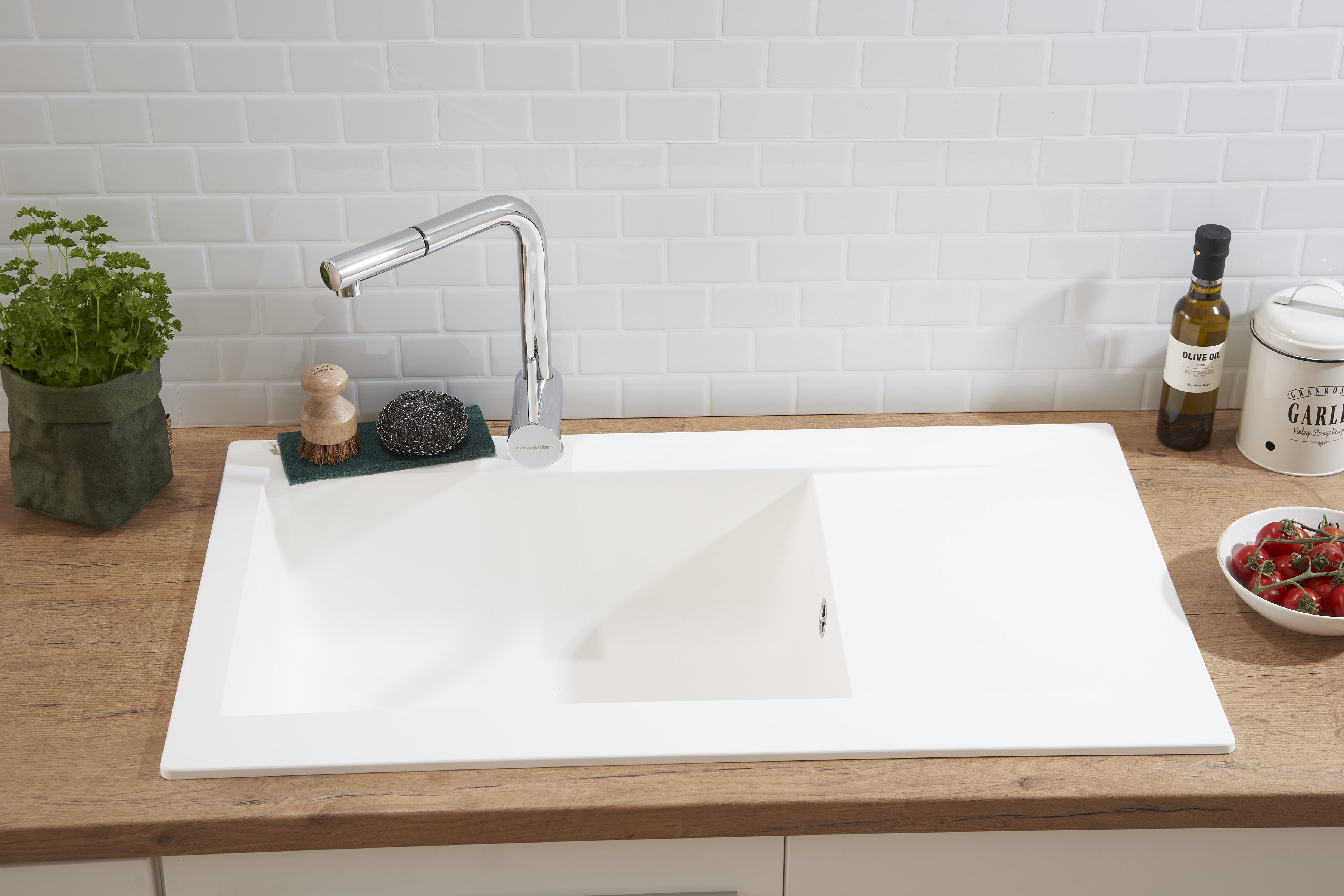 Weißes Küchenbecken mit Wasserhahn, Petersilie, Schwamm, Olivenöl und Tomaten in einem weiß gefliesten Raum.