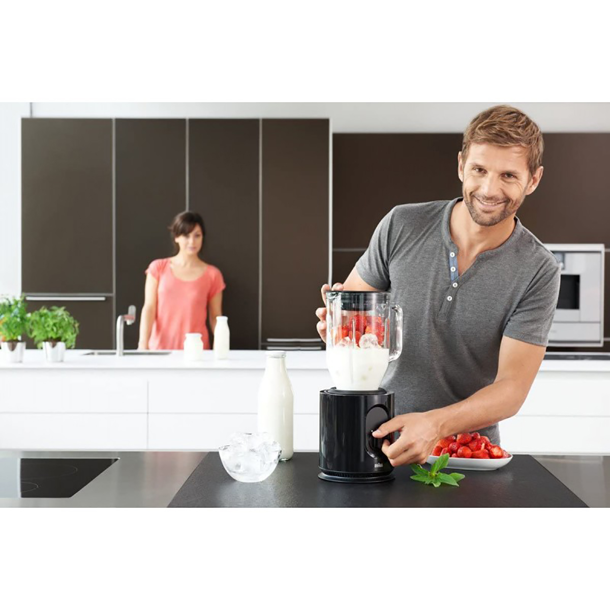 Un uomo usa un frullatore in cucina. Una donna è sullo sfondo. Ingredienti sul bancone.