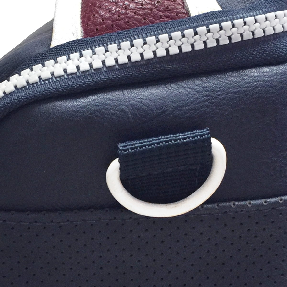 Primer plano de una bolsa azul oscuro con cremallera blanca, aro de asa blanco y detalle de cuero burdeos.