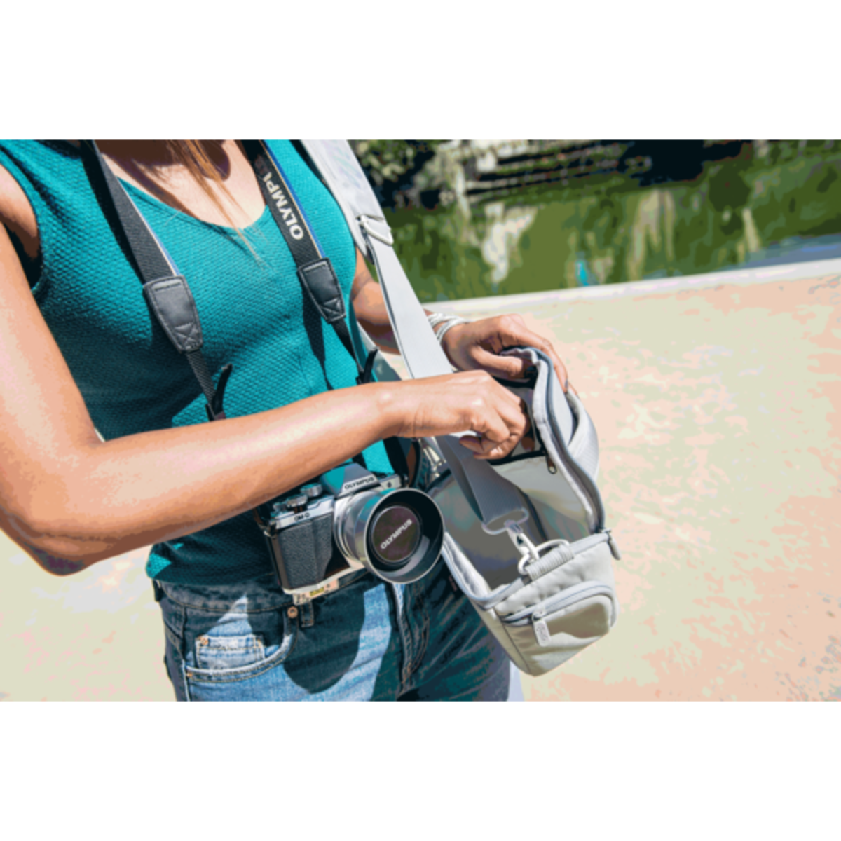 Una mujer sosteniendo una cámara. Lleva un top turquesa y vaqueros. Está abriendo una bolsa gris.