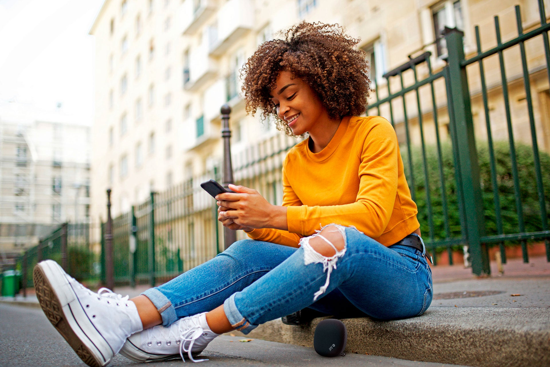 Una donna è seduta fuori, sorridendo al telefono. Indossa una camicia gialla e jeans.