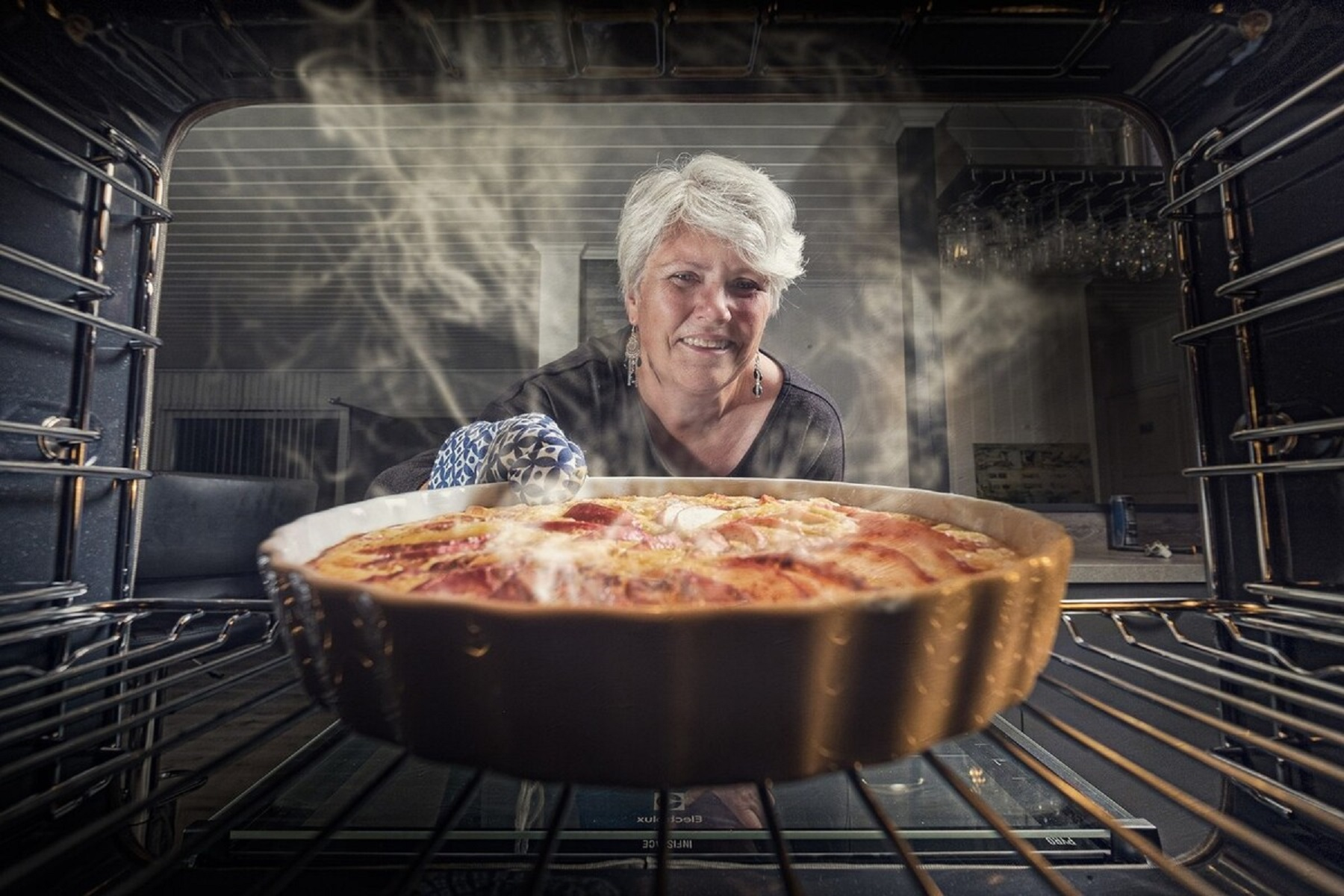 Mujer en horno, con agarradera, sonríe. Bandeja para hornear con comida dentro del horno.