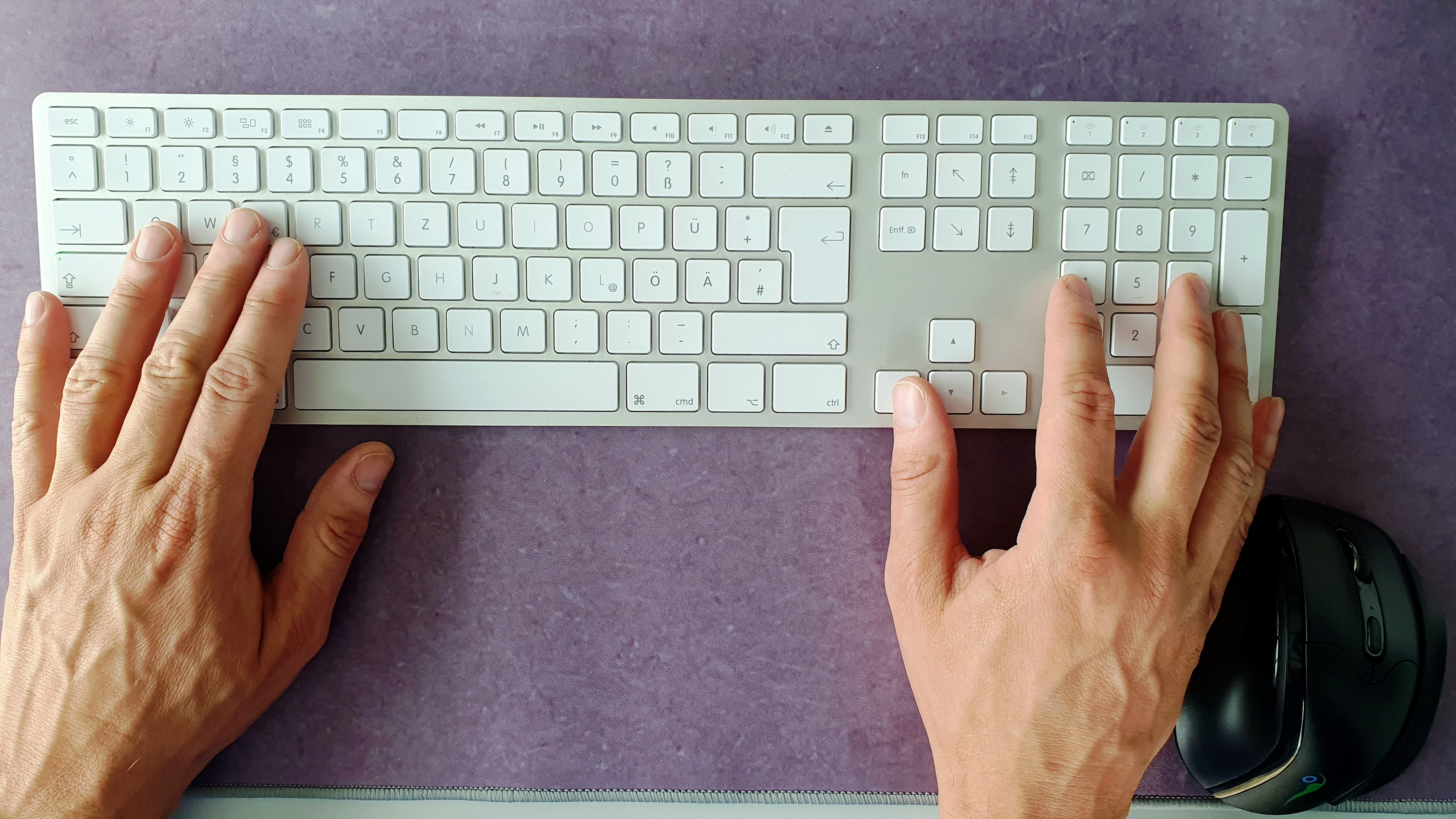 Hände tippen auf einer weißen Tastatur. Es gibt eine Maus und ein lilafarbenes Mauspad.