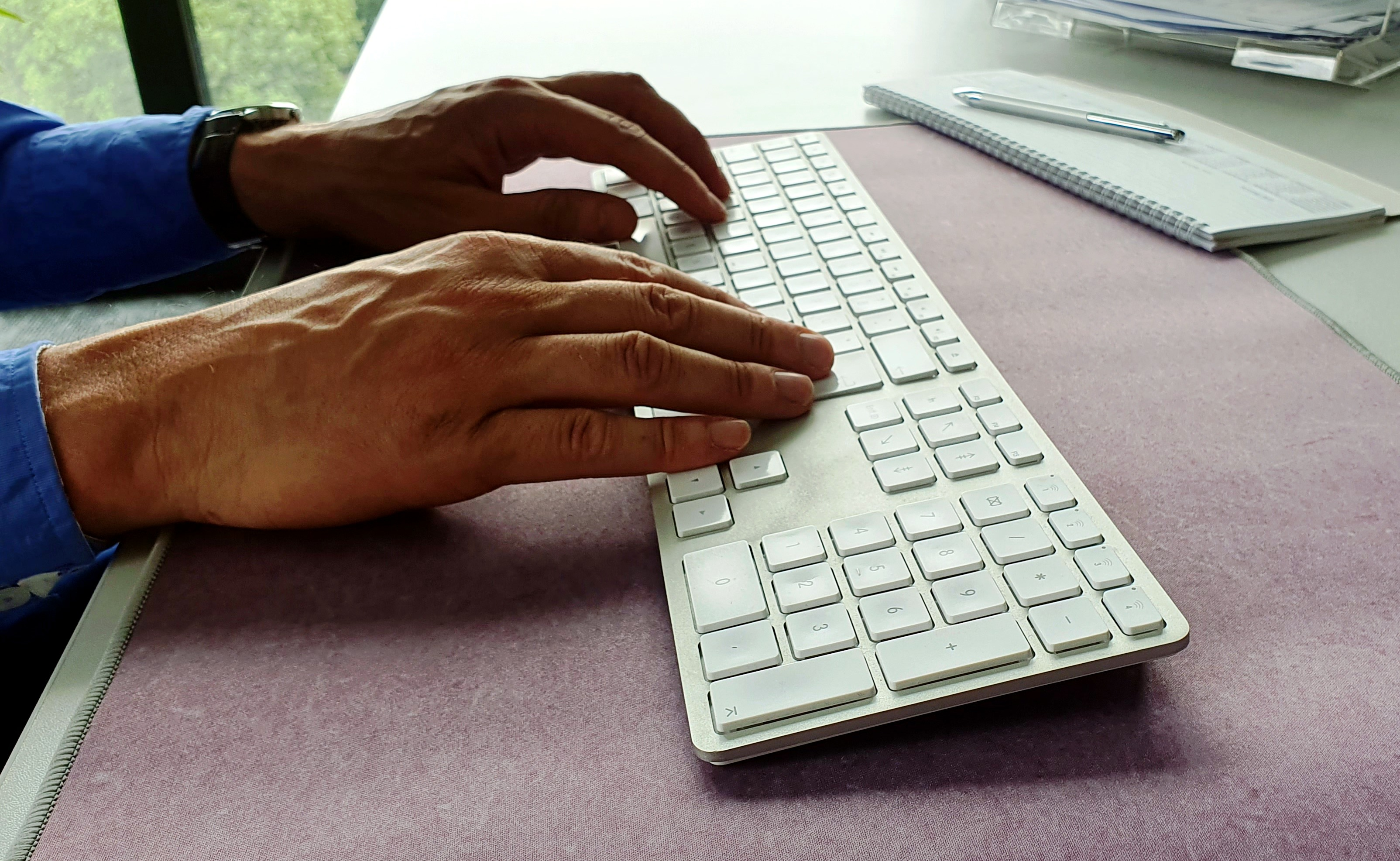 Hände tippen auf weißer Tastatur, lilafarbene Schreibtischunterlage, Notizblock.