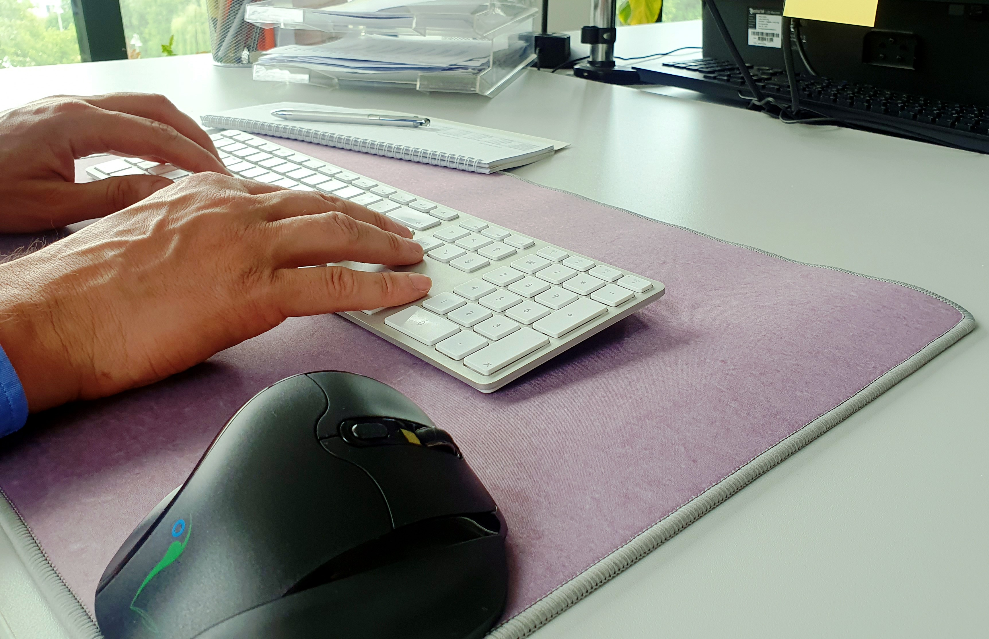 Hände tippen auf einer weißen Tastatur. Es gibt eine Maus und ein lilafarbenes Mauspad.