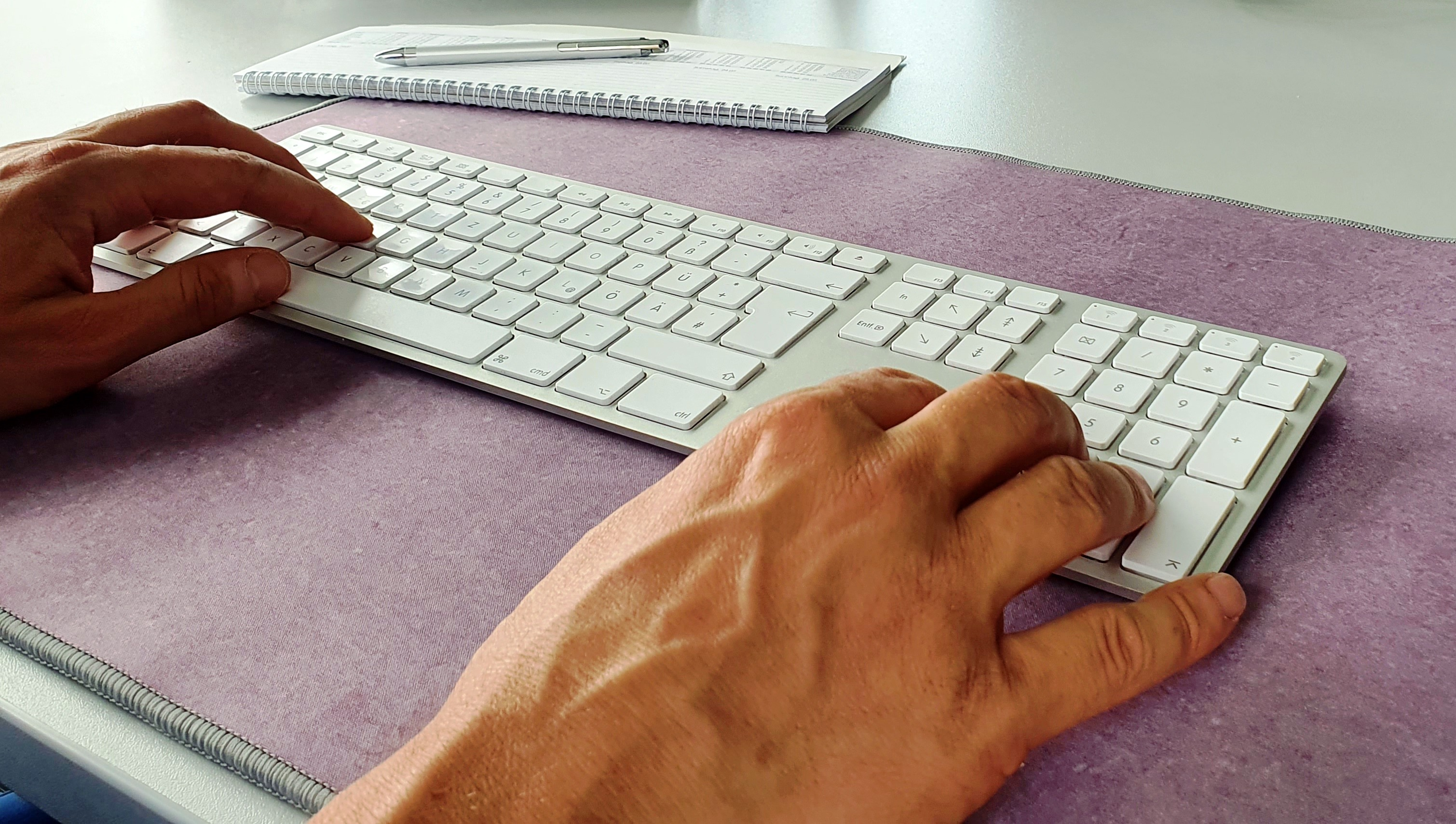 Hände, die auf einer weißen Tastatur auf einer lila Matte tippen. Ein Notizbuch und Stift sind im Hintergrund.