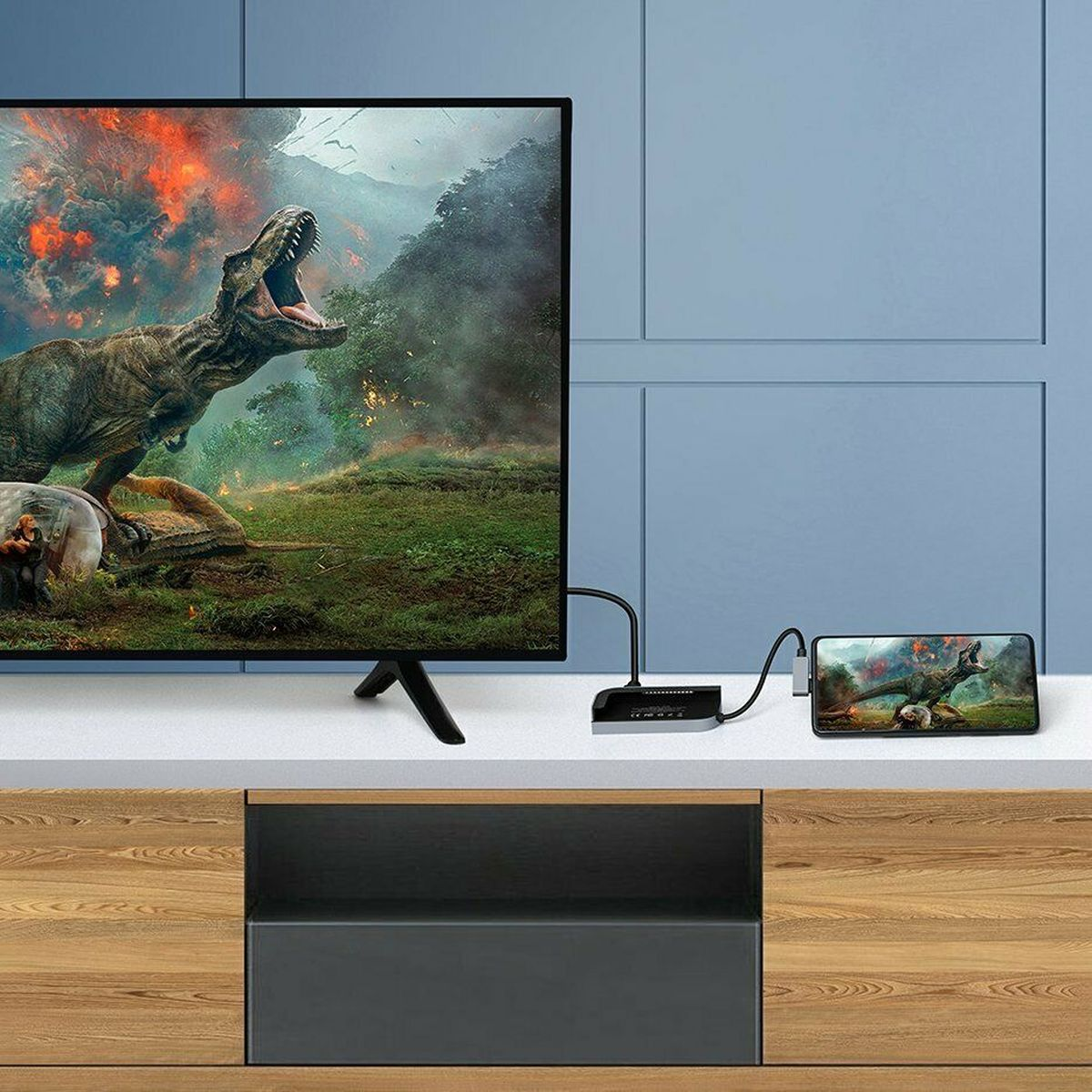TV zeigt eine Dinosaurierszene. Telefon zeigt gleiche Szene. Schwarzes Gerät und Kabel auf Tisch.