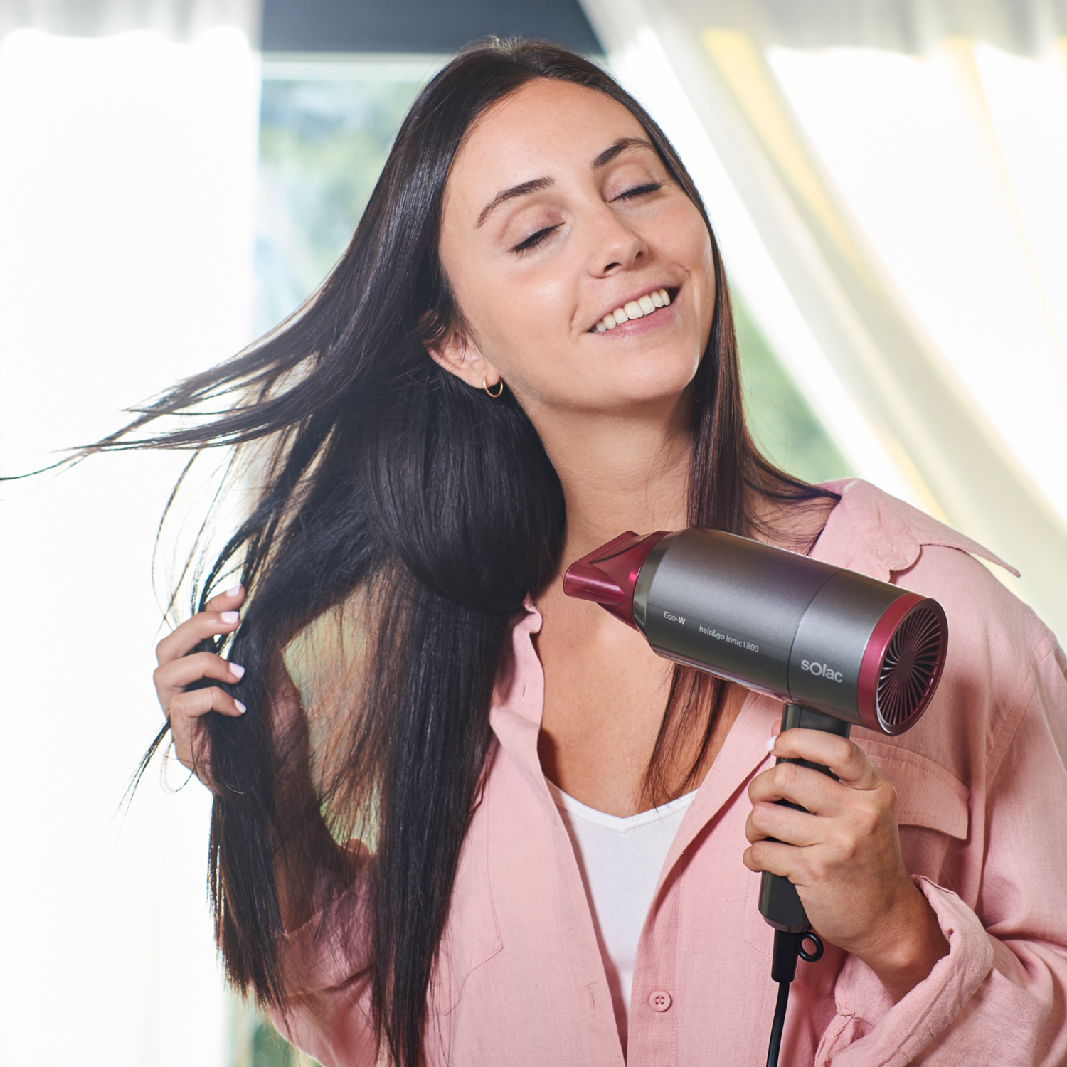 Mujer sonriendo mientras se seca el pelo con un secador. Lleva una camisa rosa.