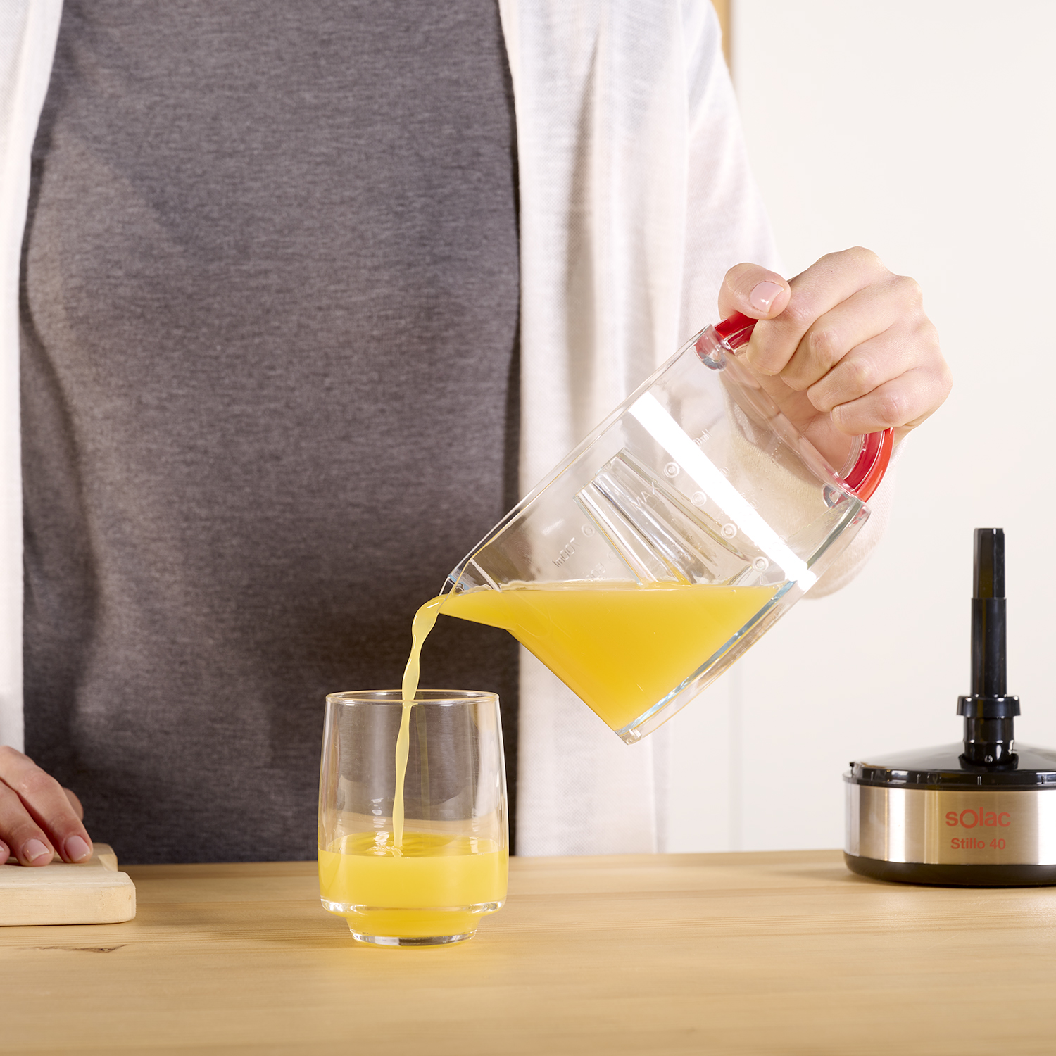 Una persona vertiendo jugo de naranja de una jarra de vidrio a un vaso. Una licuadora está en el fondo.