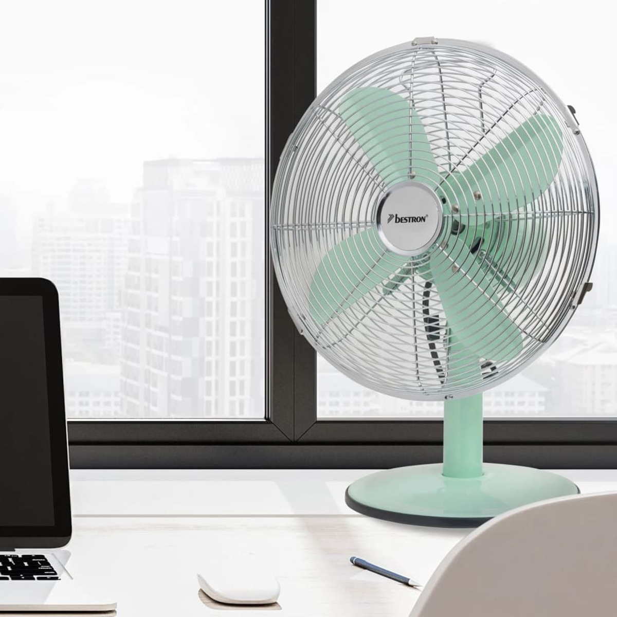 Un ventilatore da scrivania verde menta in stile retrò con gabbia e base cromate, contro una finestra e un paesaggio urbano.