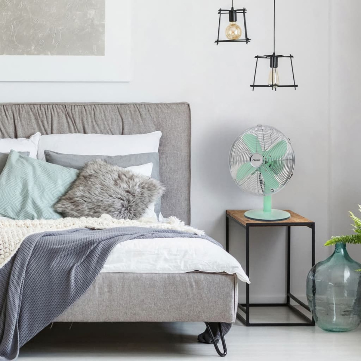 Un ventilatore verde su un tavolo vicino a un letto con cuscini e una coperta grigia.