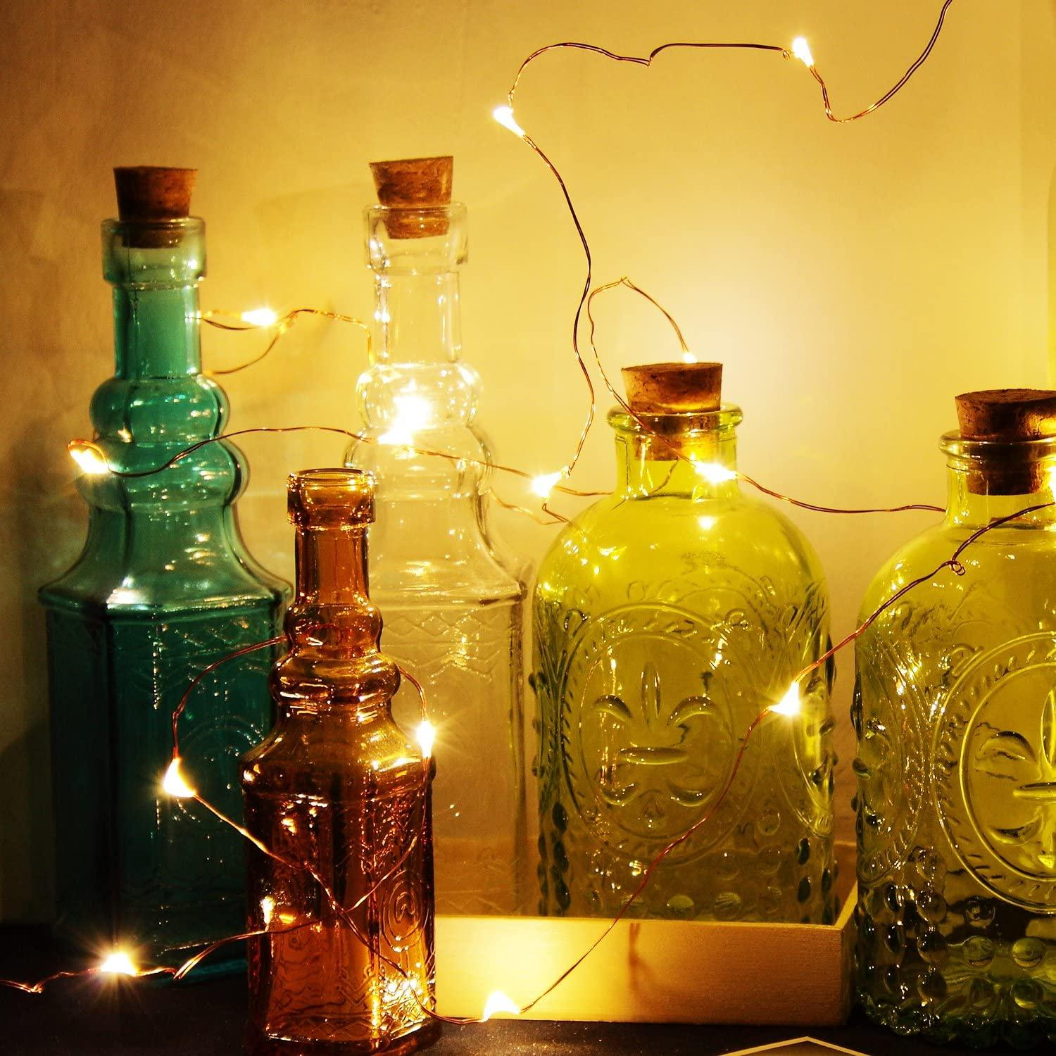 Kolekcja szklanych butelek z korkami i lampkami choinkowymi. Butelki są w różnych kolorach i kształtach.