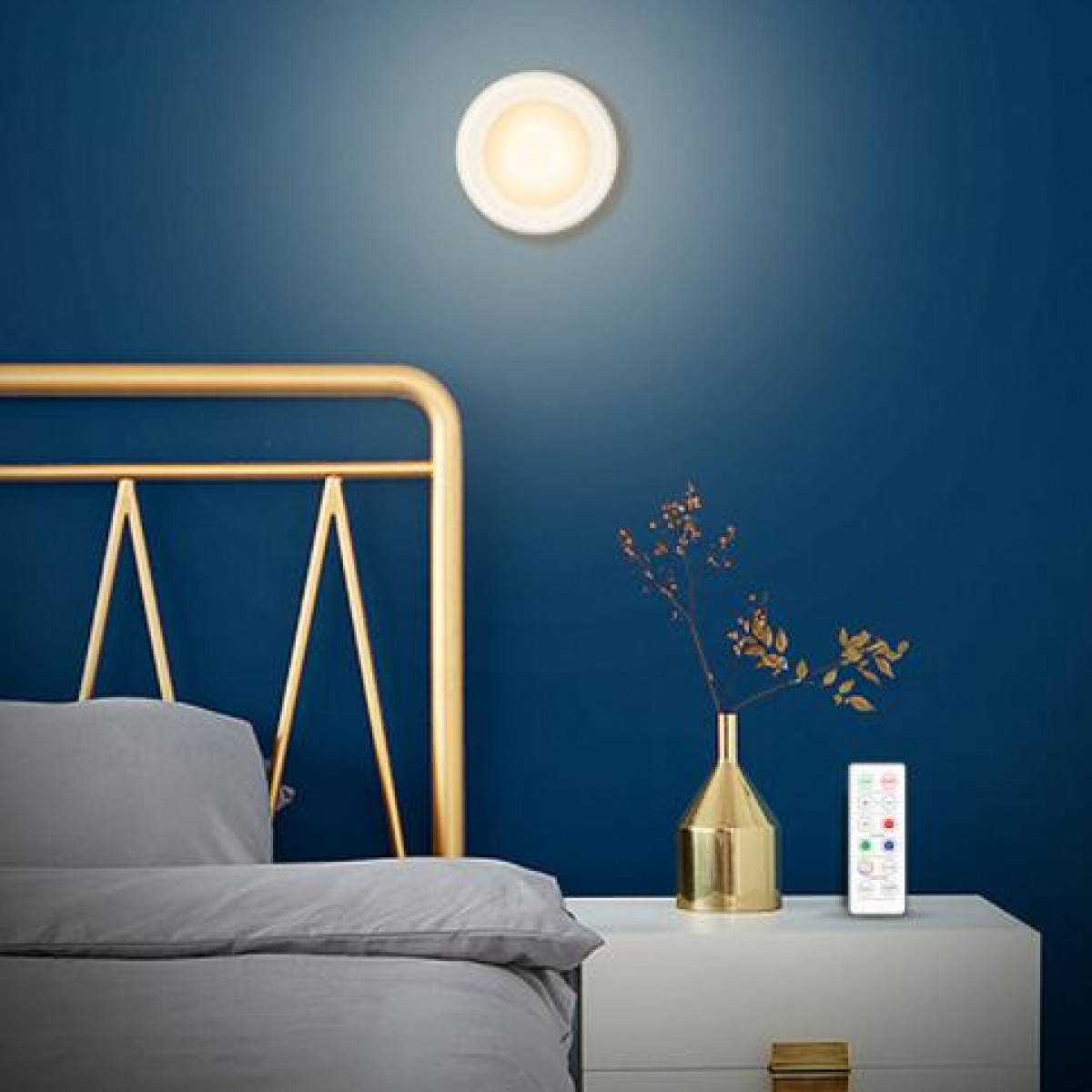 Una luz circular montada en la pared con un mando a distancia en una mesita de noche blanca. Un marco de cama dorado.