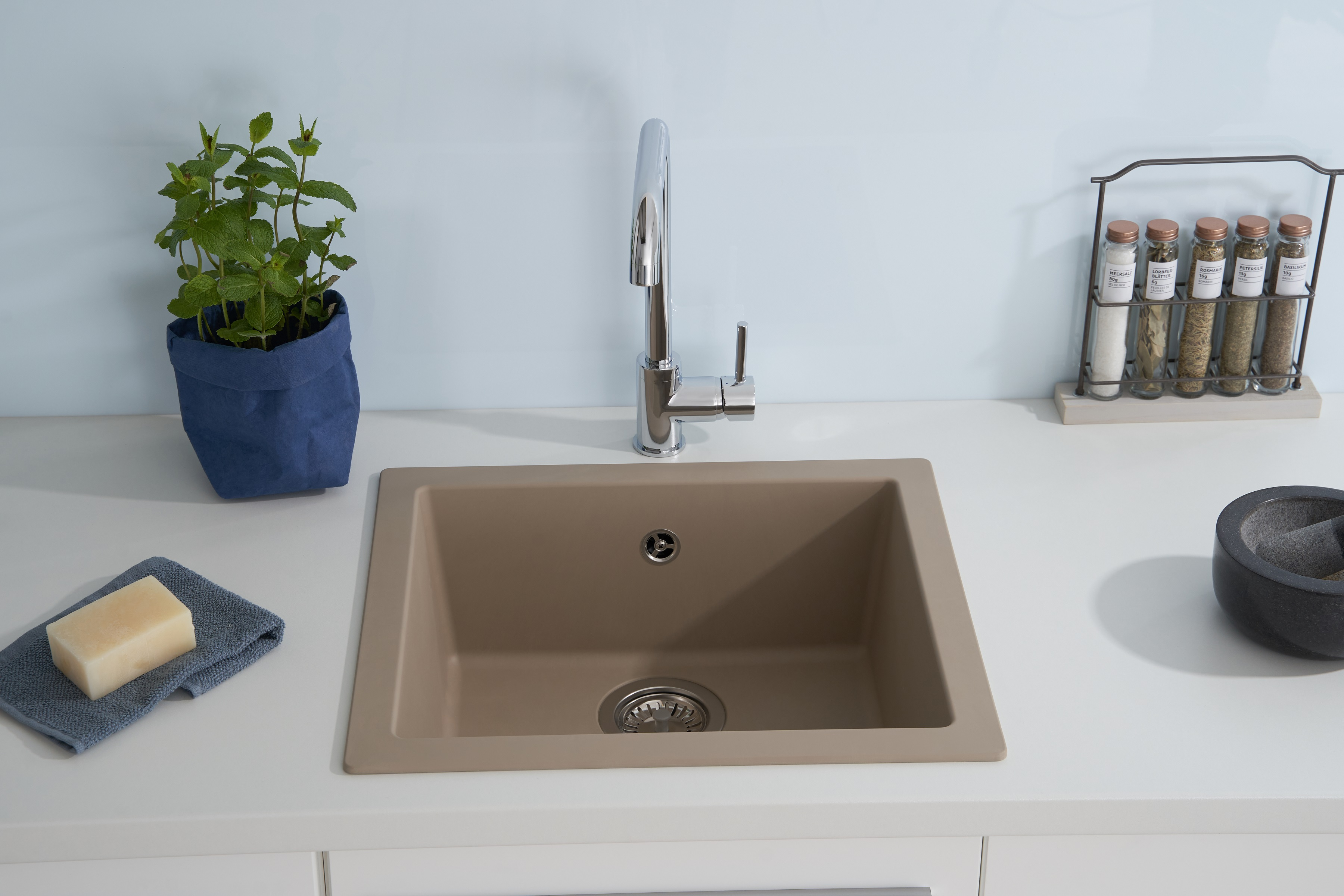 Beiges Küchenbecken mit Chromarmatur, Seife und Pflanze in blauem Topf auf weißer Arbeitsplatte.
