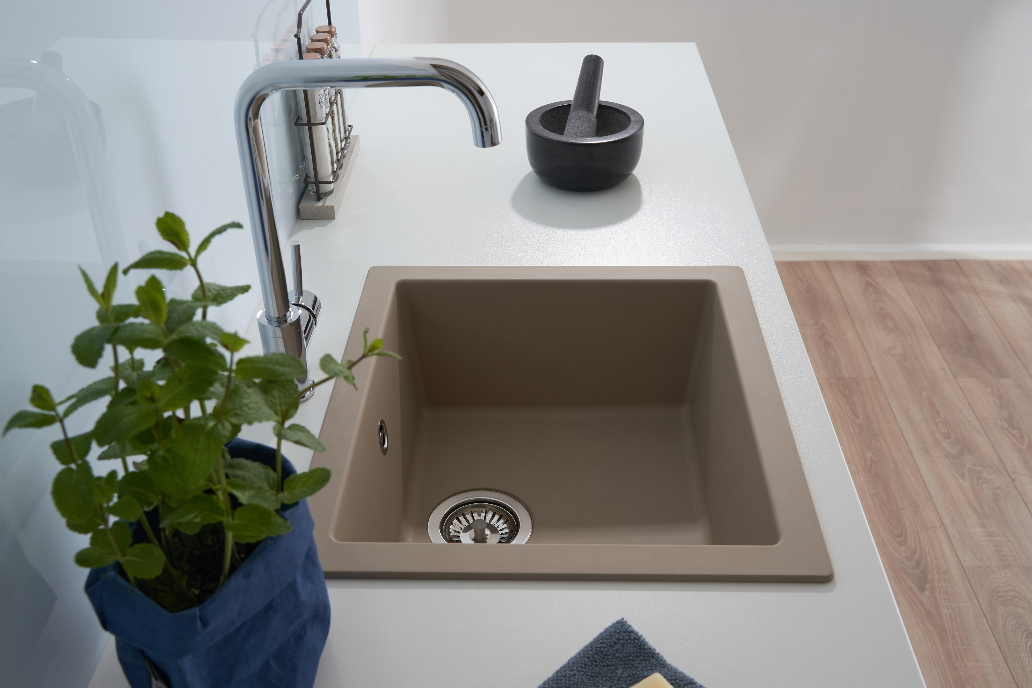 Beiges Küchenbecken mit Wasserhahn, Pflanze davor, Mörser rechts, weiße Arbeitsplatte.