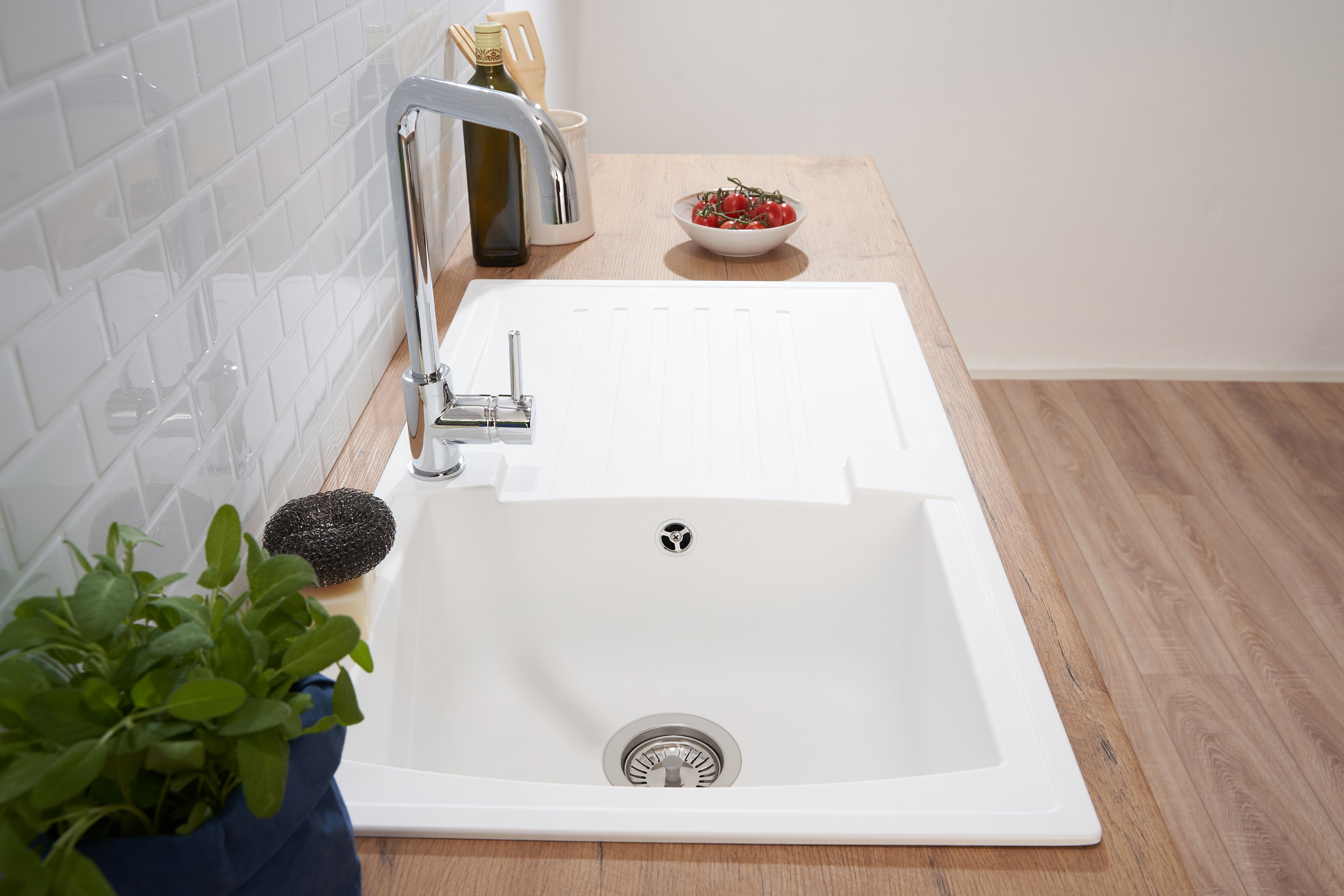 Weißes Küchenwaschbecken mit Wasserhahn. Gemüse und Ölflasche an der Seite. Weiße Fliesen im Hintergrund.