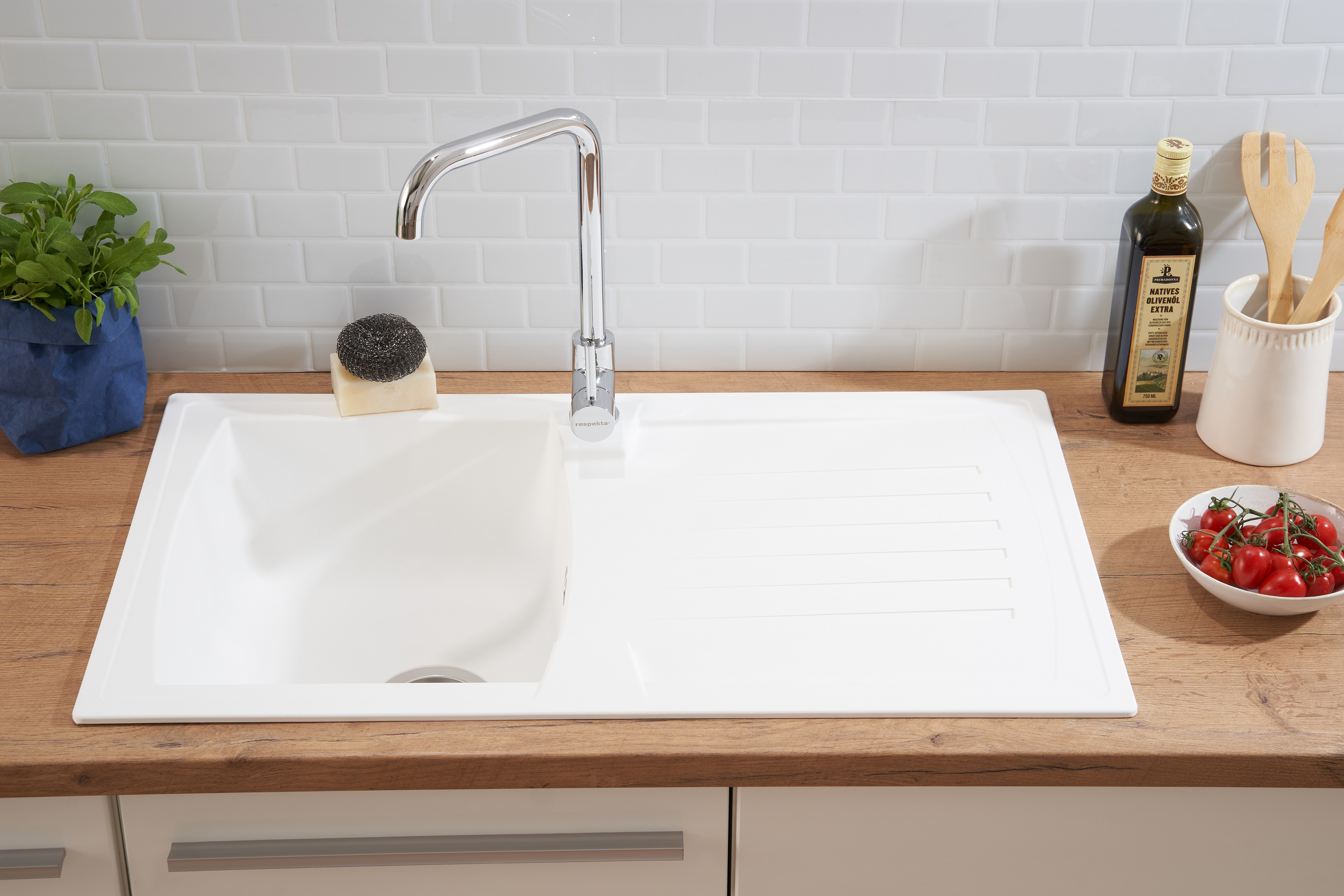 Ein Küchenspülbecken mit Wasserhahn, Tomaten und Olivenöl. Das Becken ist weiß, die Arbeitsplatte Holz.