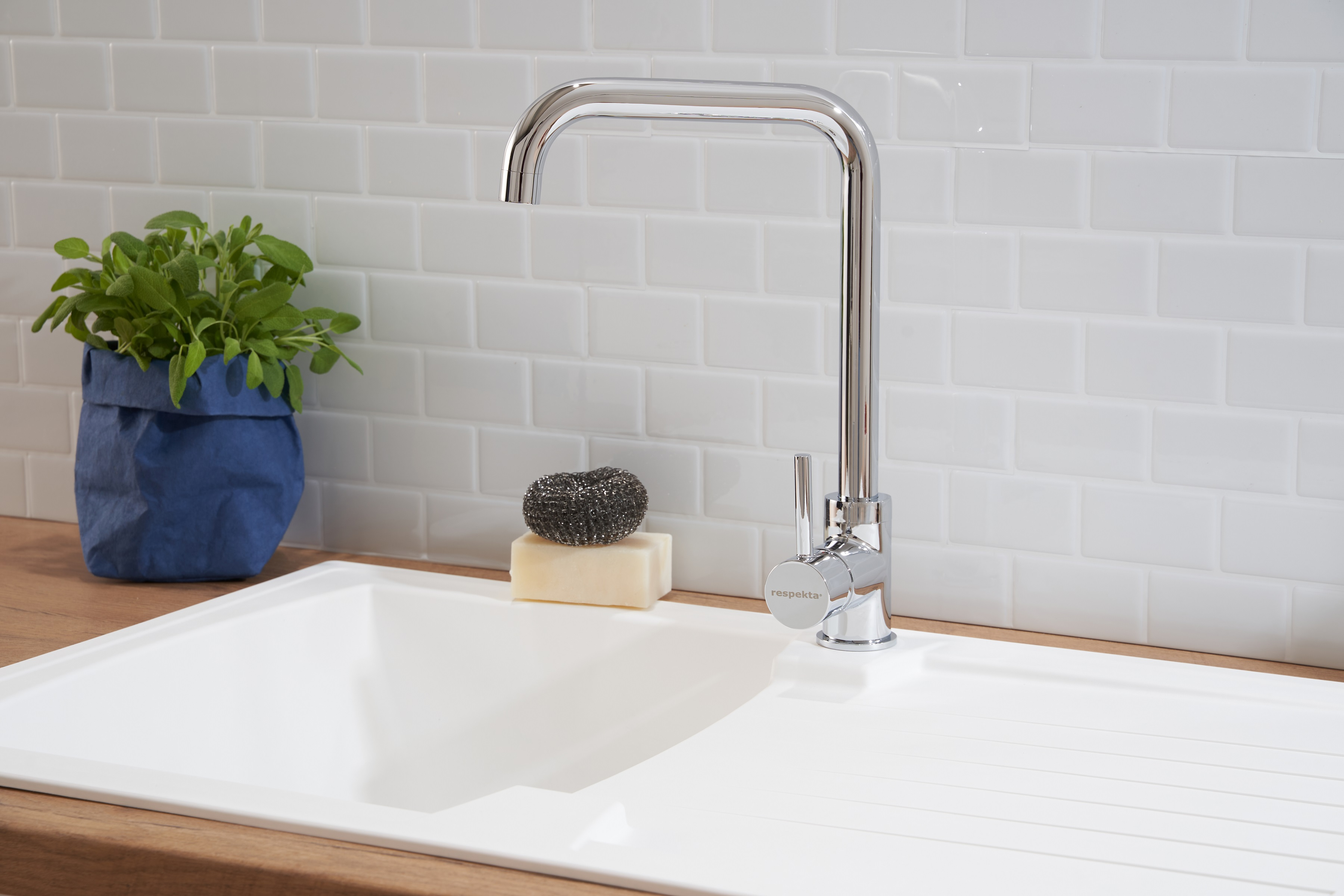 Küchenspüle mit Wasserhahn, weiße Fliesen, Seife und eine Pflanze. Helle und saubere Umgebung.