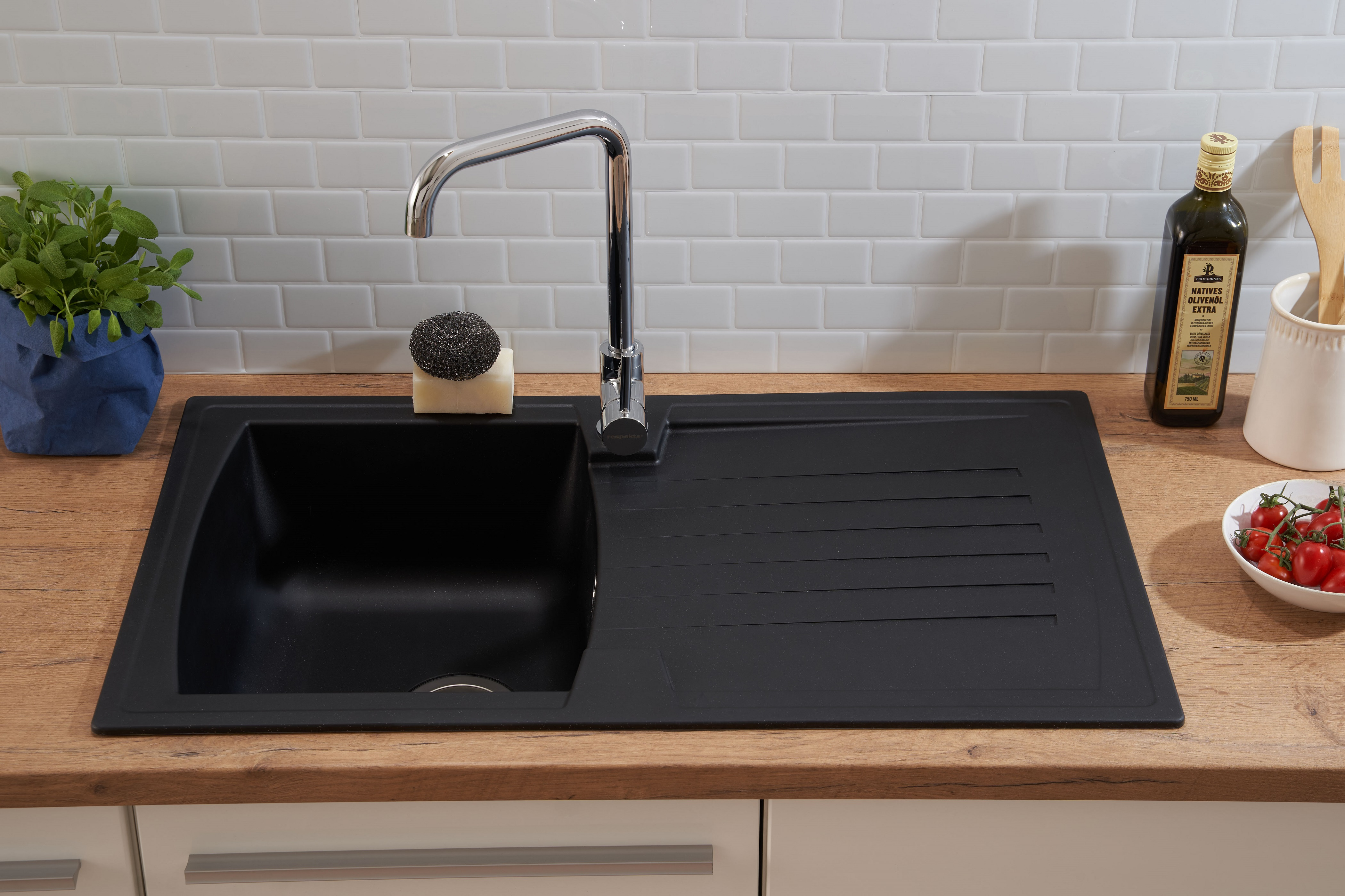 Schwarzes Küchenbecken mit Chromarmatur, Schwamm, Olivenöl und Tomaten auf Holz-Arbeitsplatte.
