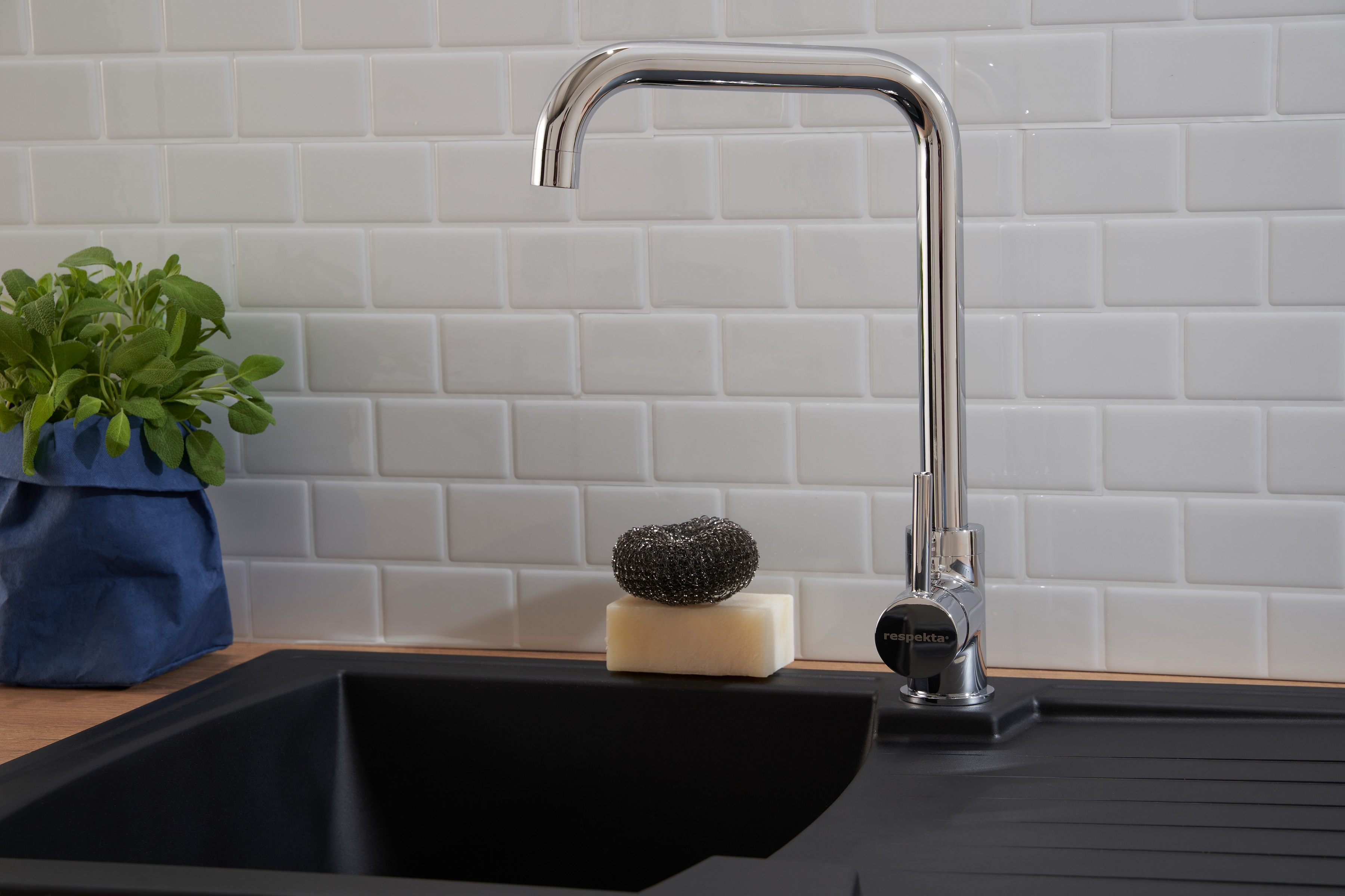 Eine Chromarmatur über einem schwarzen Waschbecken. Seife, Schwamm und Pflanze stehen auf der Theke.
